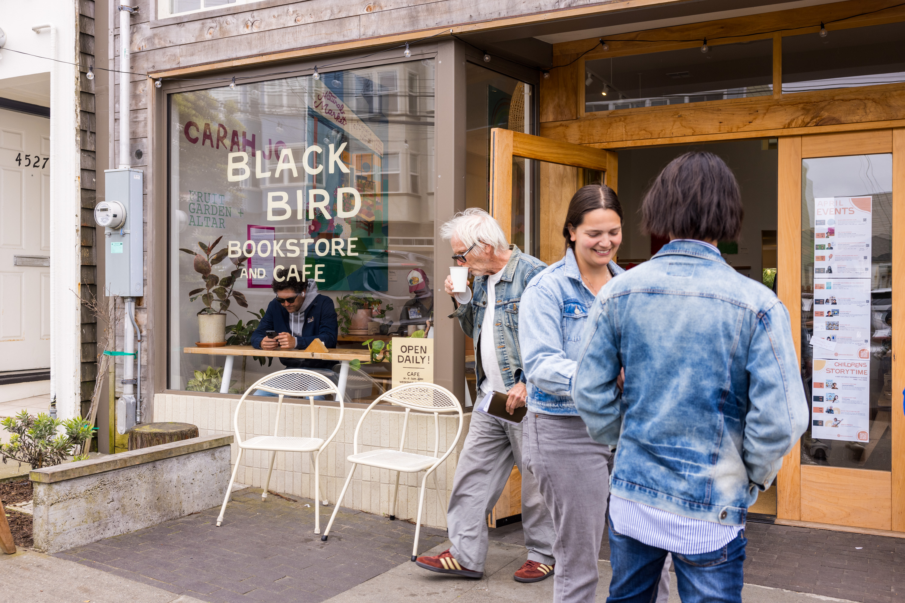 A group of people interact outside Black Bird Bookstore and Cafe. One person is seated inside on a phone, while another enjoys a drink. The ambiance is casual and welcoming.