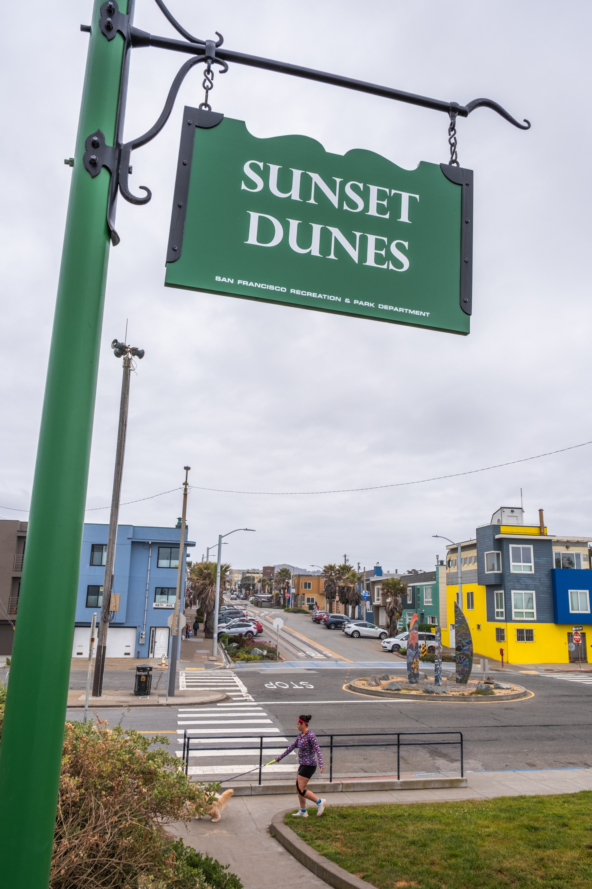 A green sign reads &quot;Sunset Dunes.&quot; Below, a person walks a dog on a sidewalk in a neighborhood with colorful buildings and palm trees under a cloudy sky.