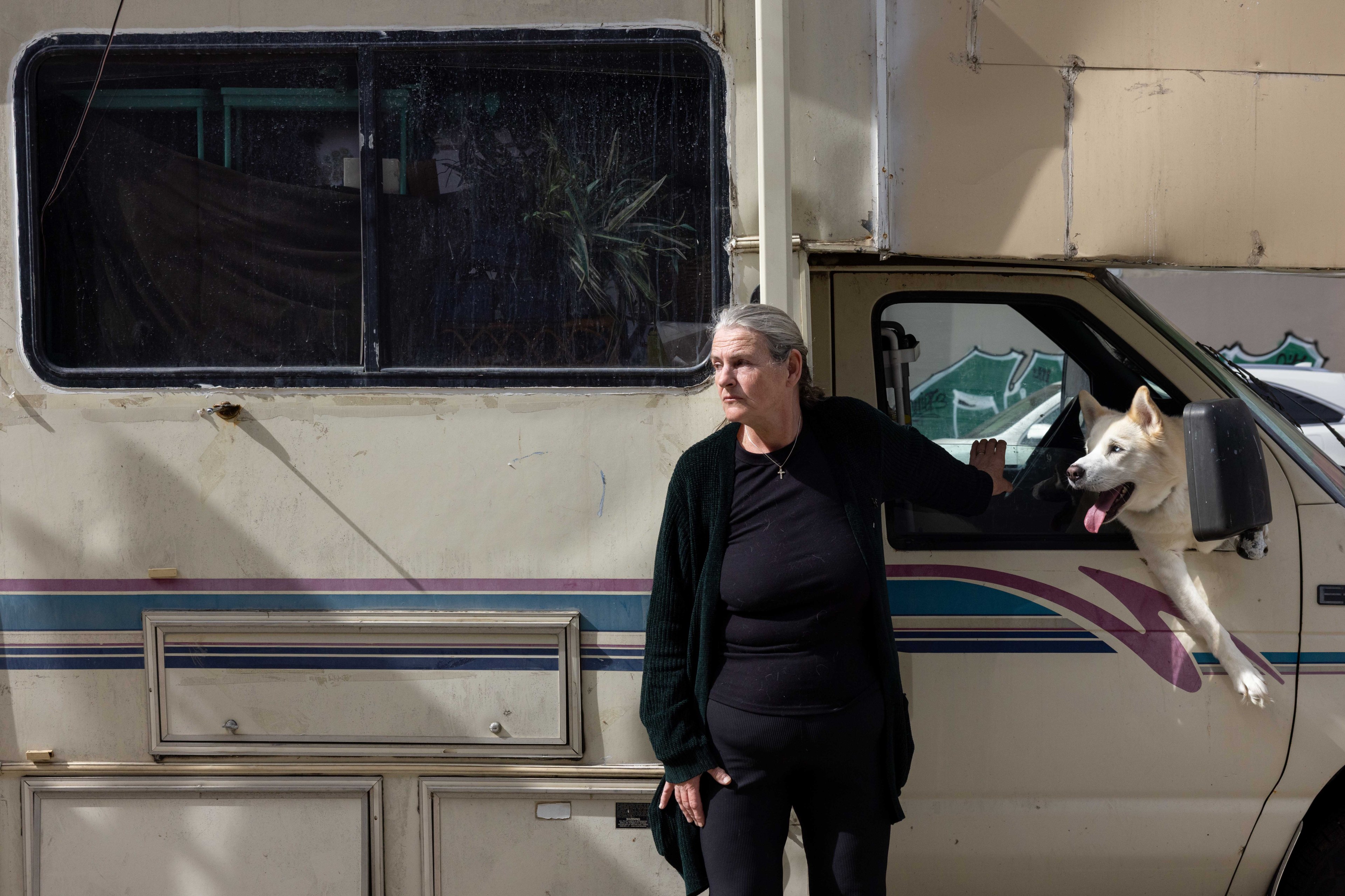 A woman in black stands leaning against an RV, with a white dog sticking its head and paws out of the driver's window. The RV is parked on a street.