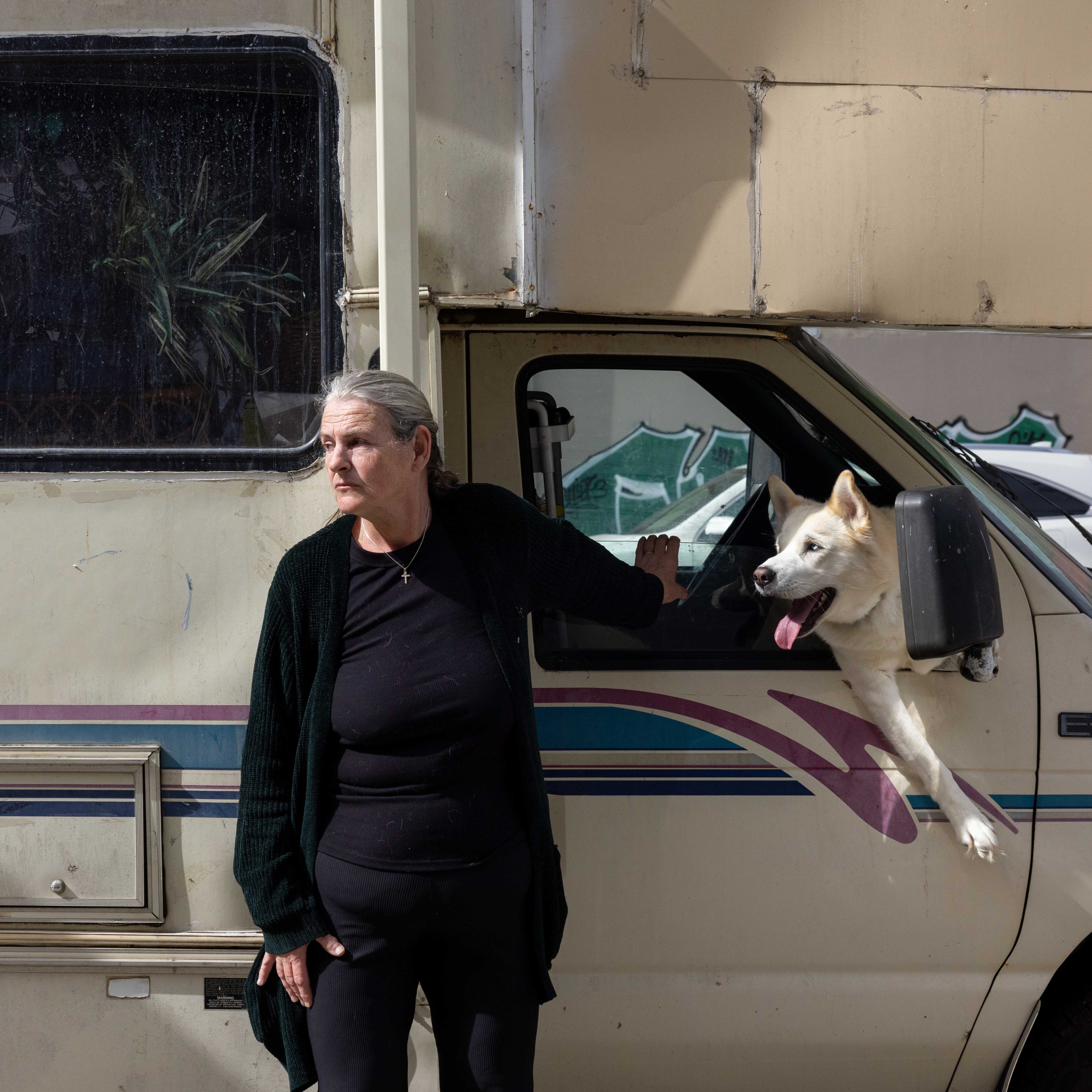 A woman in black stands leaning against an RV, with a white dog sticking its head and paws out of the driver's window. The RV is parked on a street.