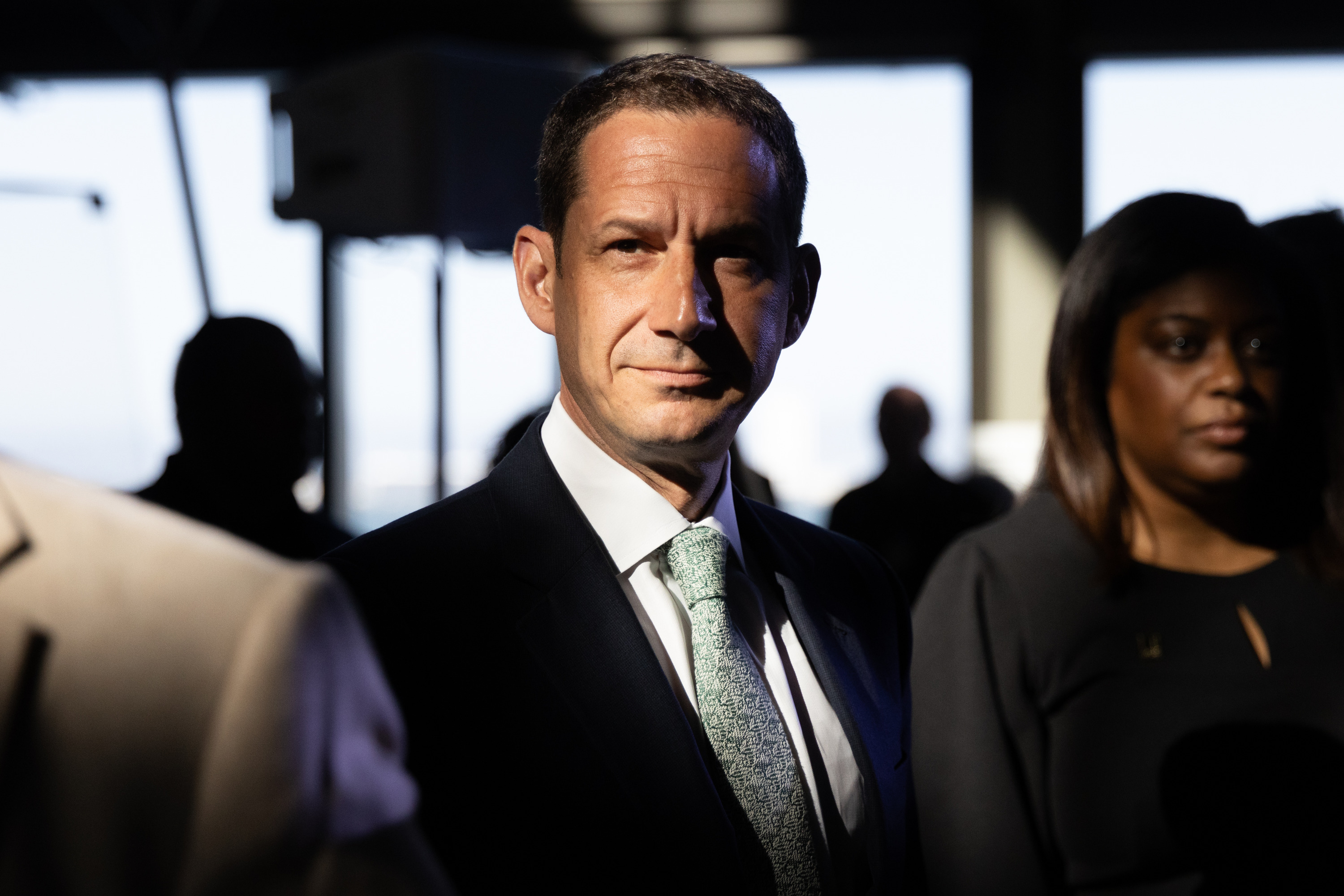 A man in a suit stands under bright lighting, creating strong contrasts. He is surrounded by people in a dimly lit room with large, bright windows and blurred figures.