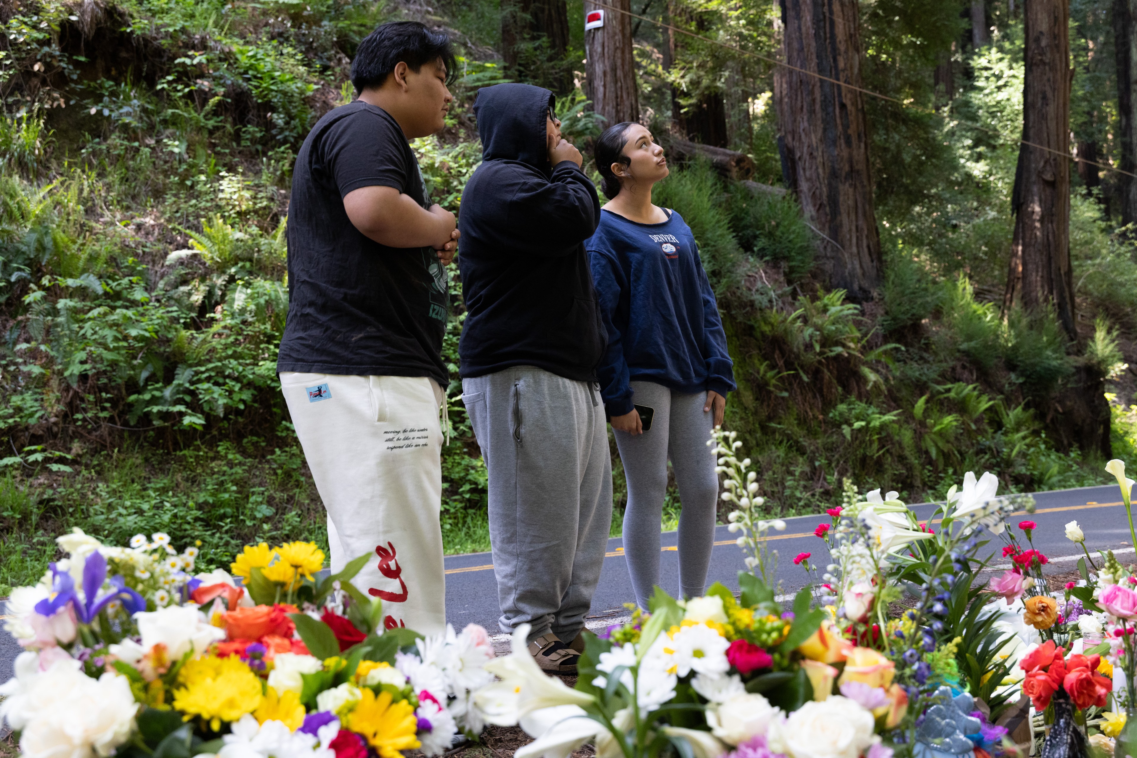 Three people stand solemnly by a roadside adorned with colorful flowers. They're surrounded by tall trees, creating a serene natural backdrop.