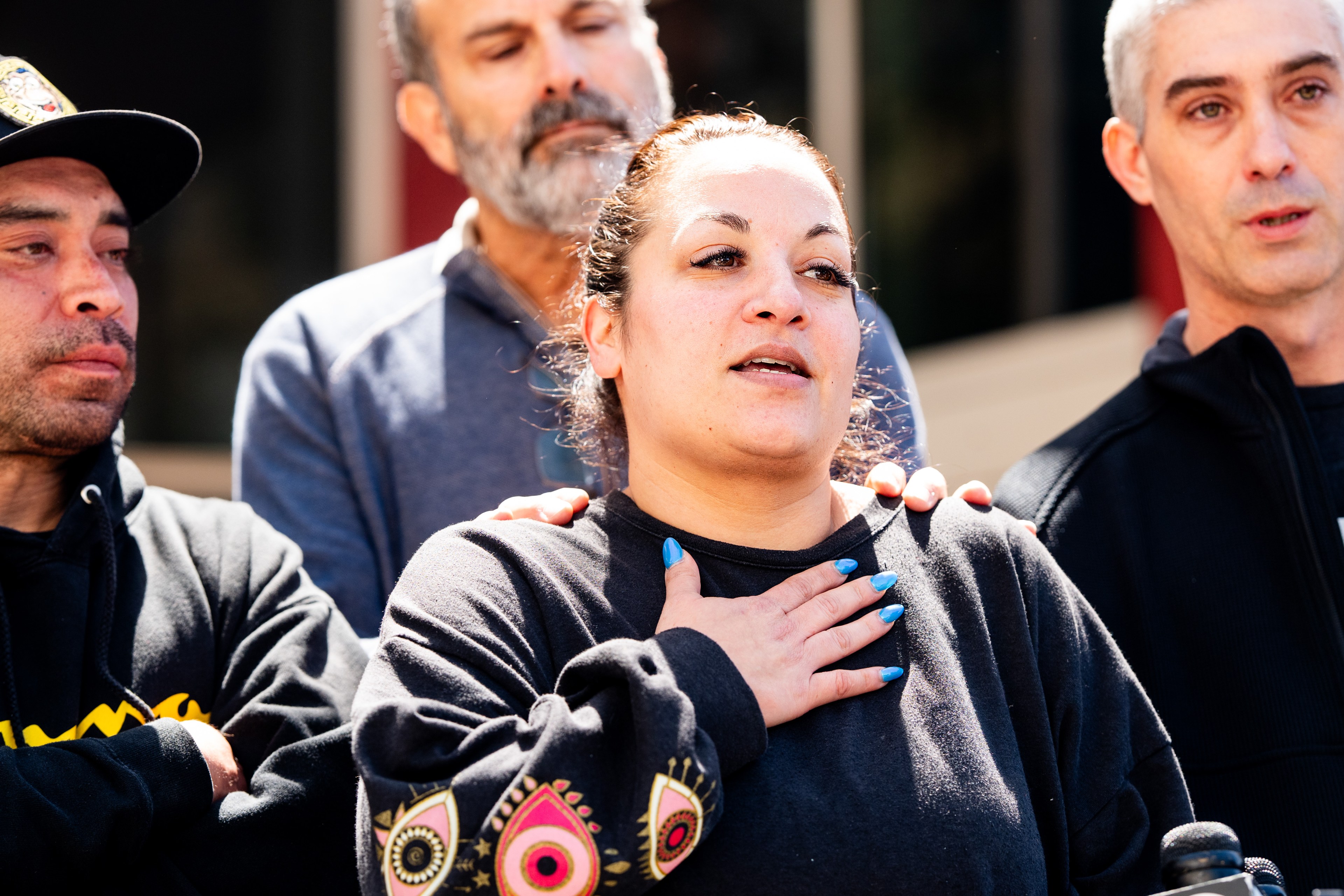 A woman stands in the center holding her chest, surrounded by three men. Two men touch her shoulders in a supportive gesture. All have solemn expressions.