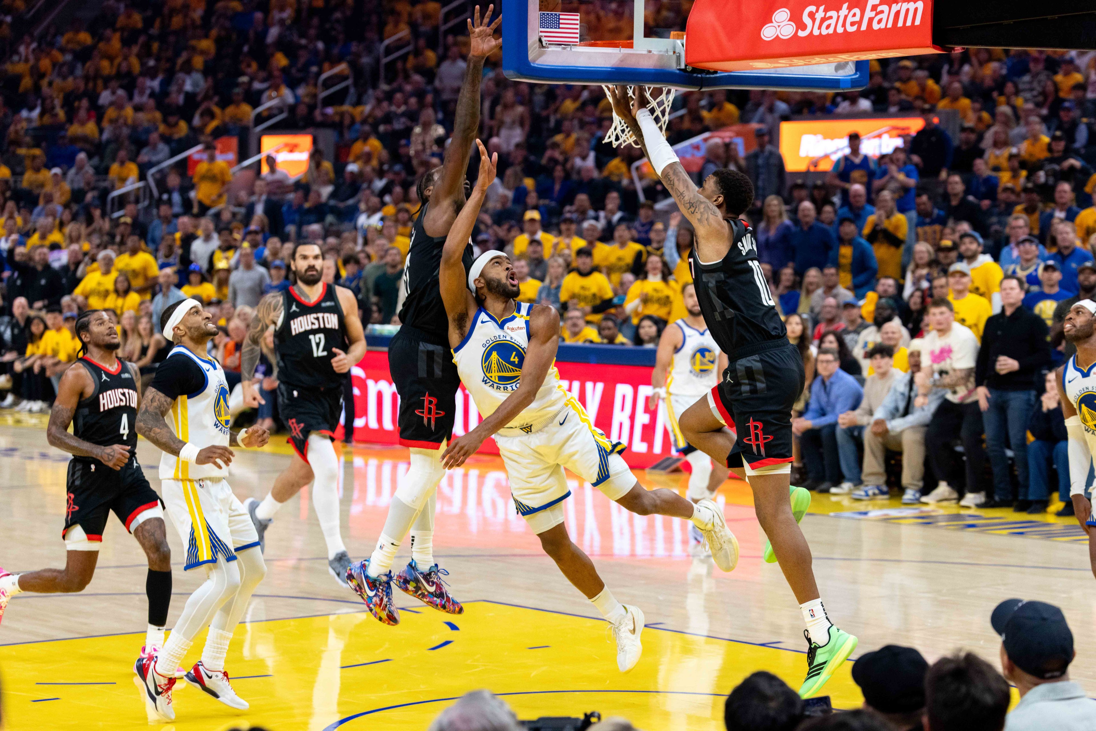 In a basketball game, a player in a black uniform jumps to make a layup while being defended by players in white and blue. The audience watches intently from the stands.