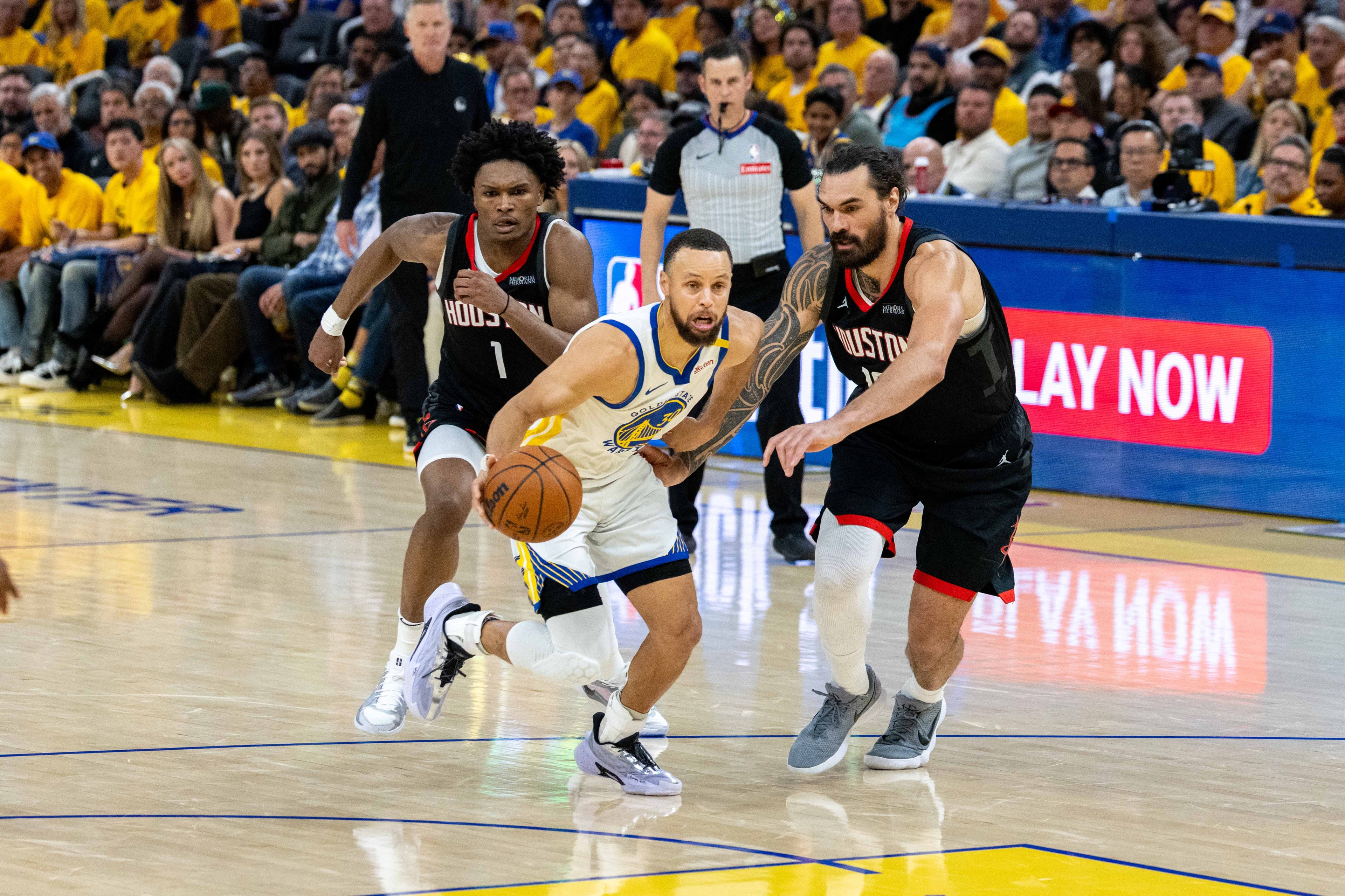 A basketball player in a Warriors uniform dribbles the ball while two opposing players in black Rockets uniforms defend him on a crowded indoor court.