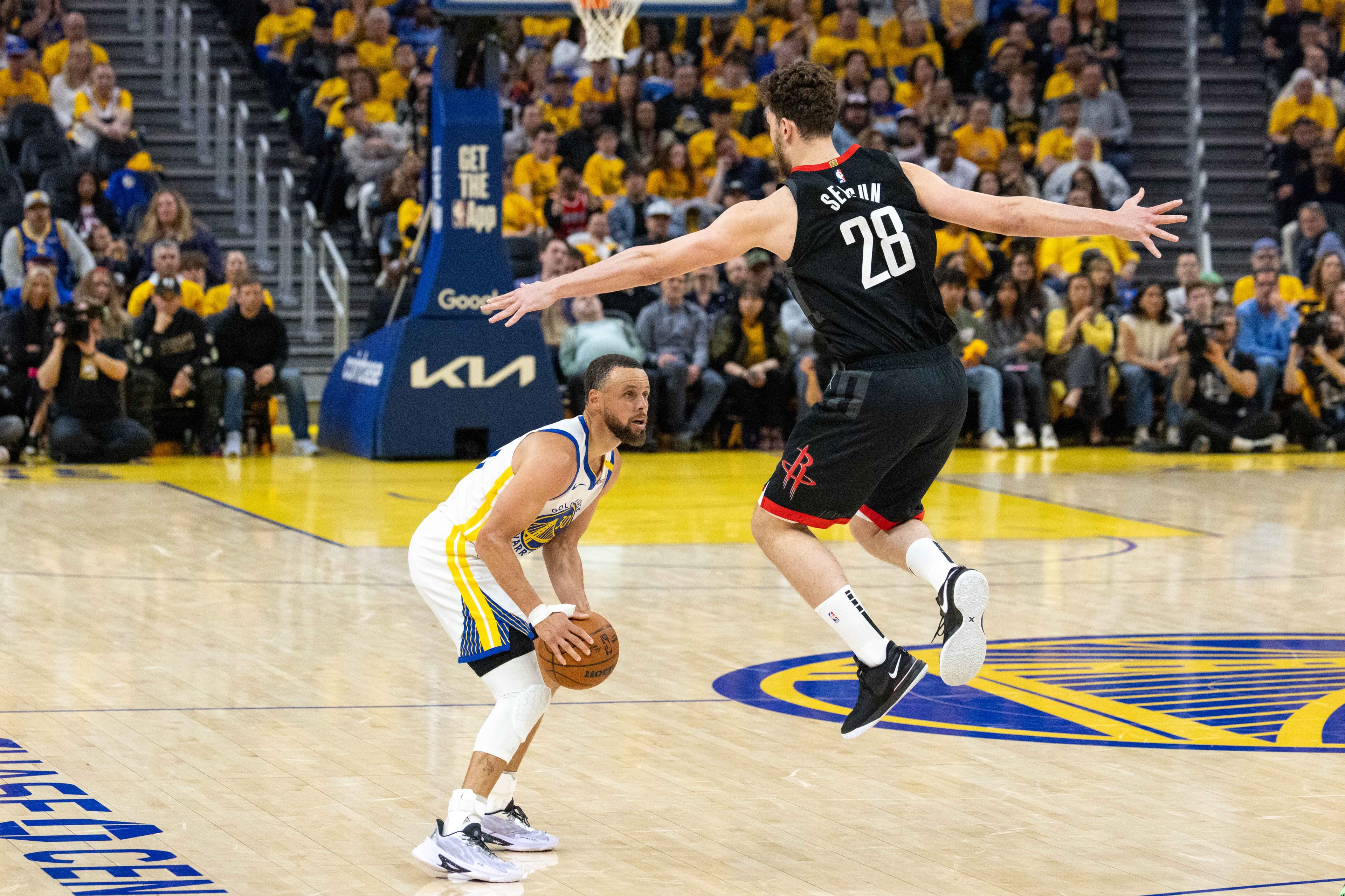 A basketball player in a yellow jersey prepares to shoot as another player in a black jersey jumps to block. The court is surrounded by spectators.
