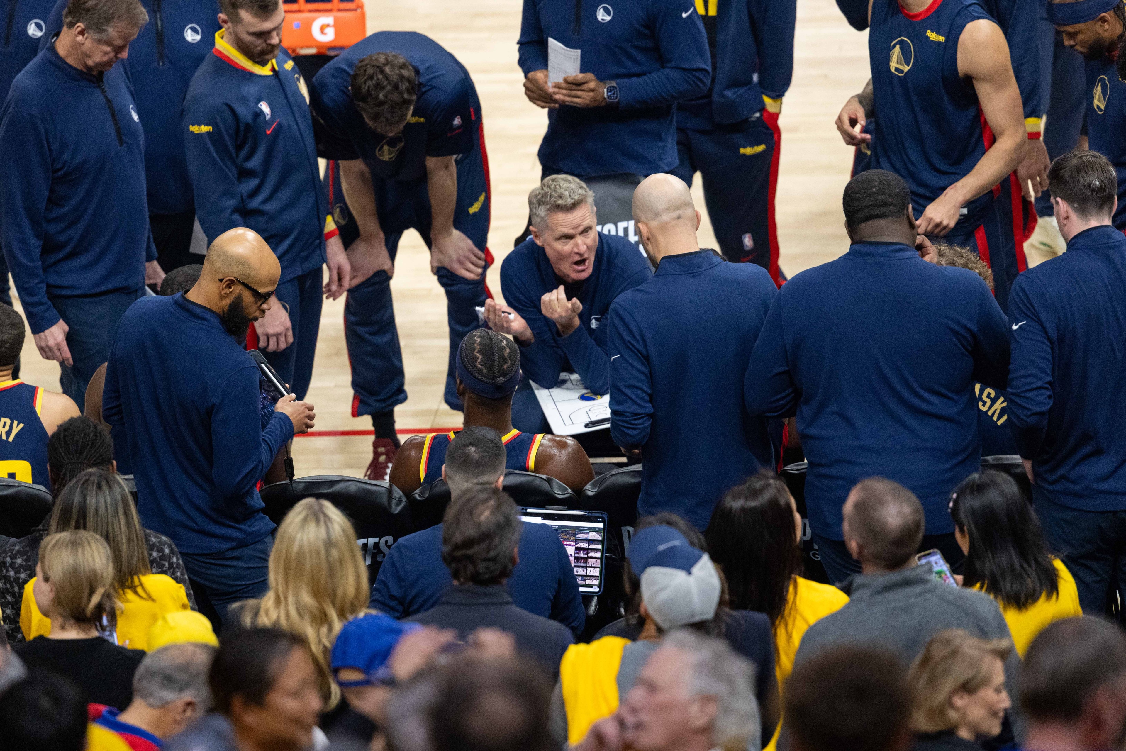 A basketball coach, surrounded by players in navy tracksuits, passionately discusses tactics with a seated player during a timeout, while onlookers watch attentively.