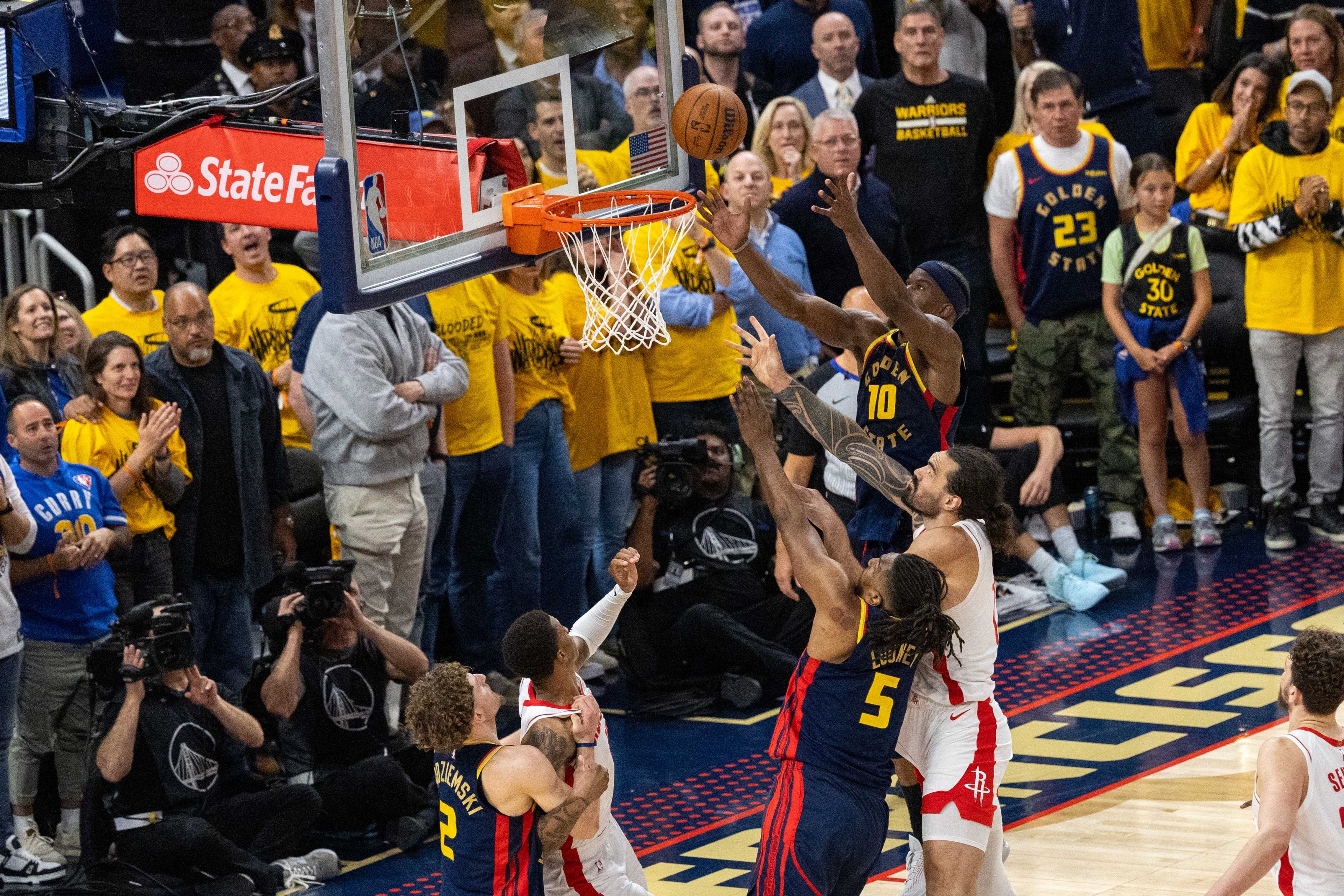 A basketball game with a player in a blue "Golden State" jersey attempting a shot near the hoop. Fans in yellow shirts watch, while cameras capture the action.