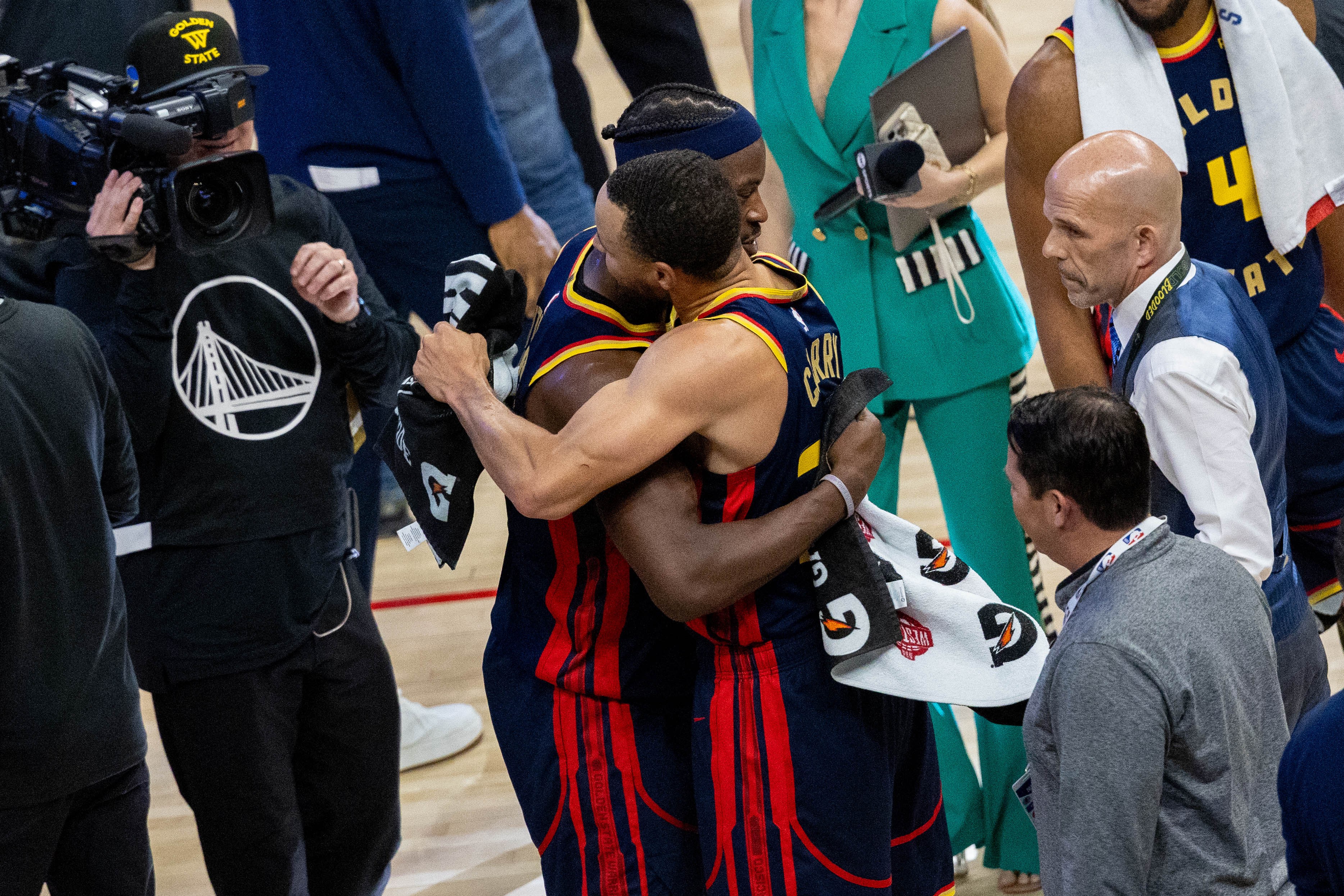 Two basketball players in red and blue jerseys hug surrounded by media and staff. A cameraman captures the moment as a woman in a green outfit stands nearby.