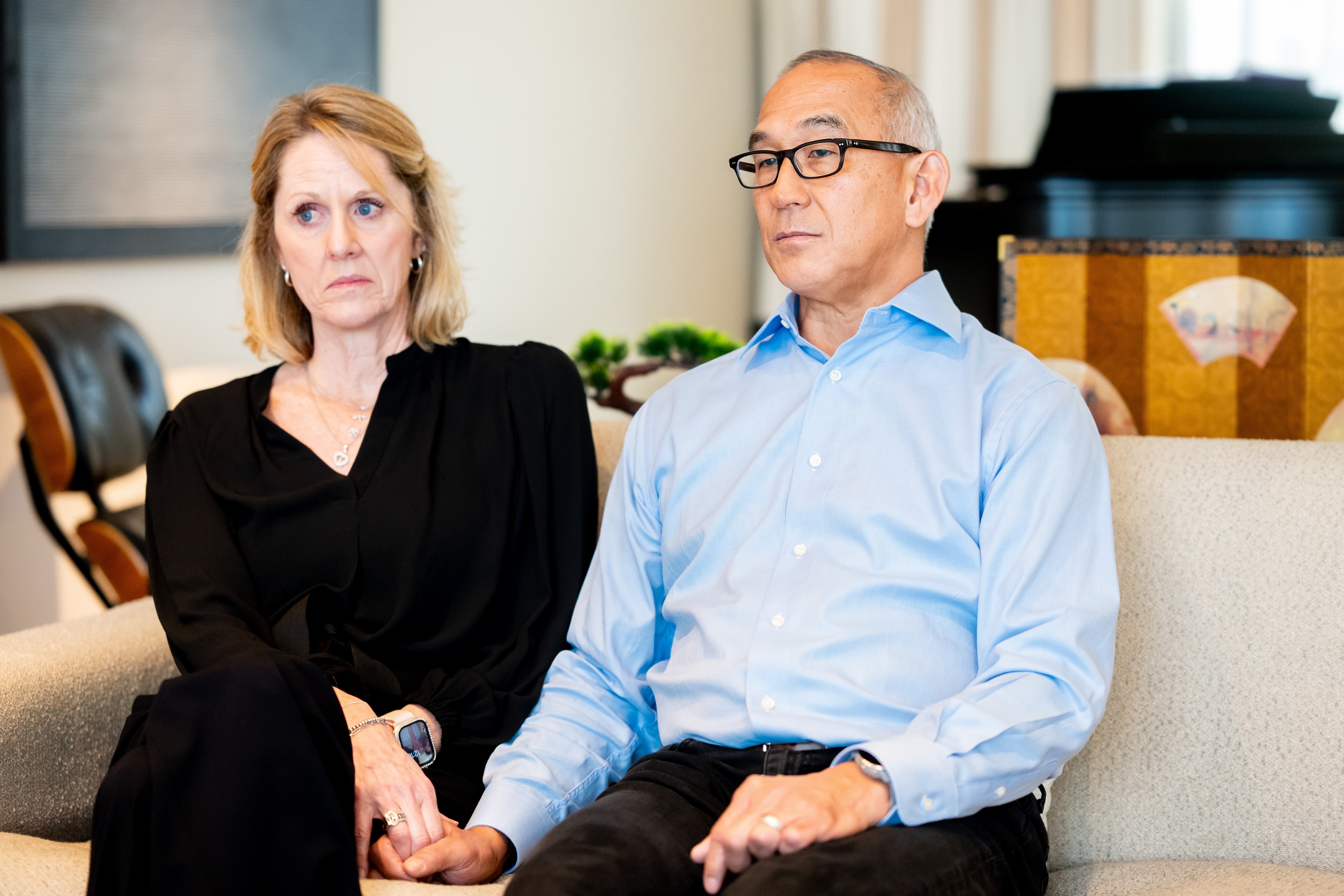 A man and woman sit closely on a sofa, holding hands. The woman wears black, and the man is in a blue shirt. Both have neutral expressions, in a cozy room.