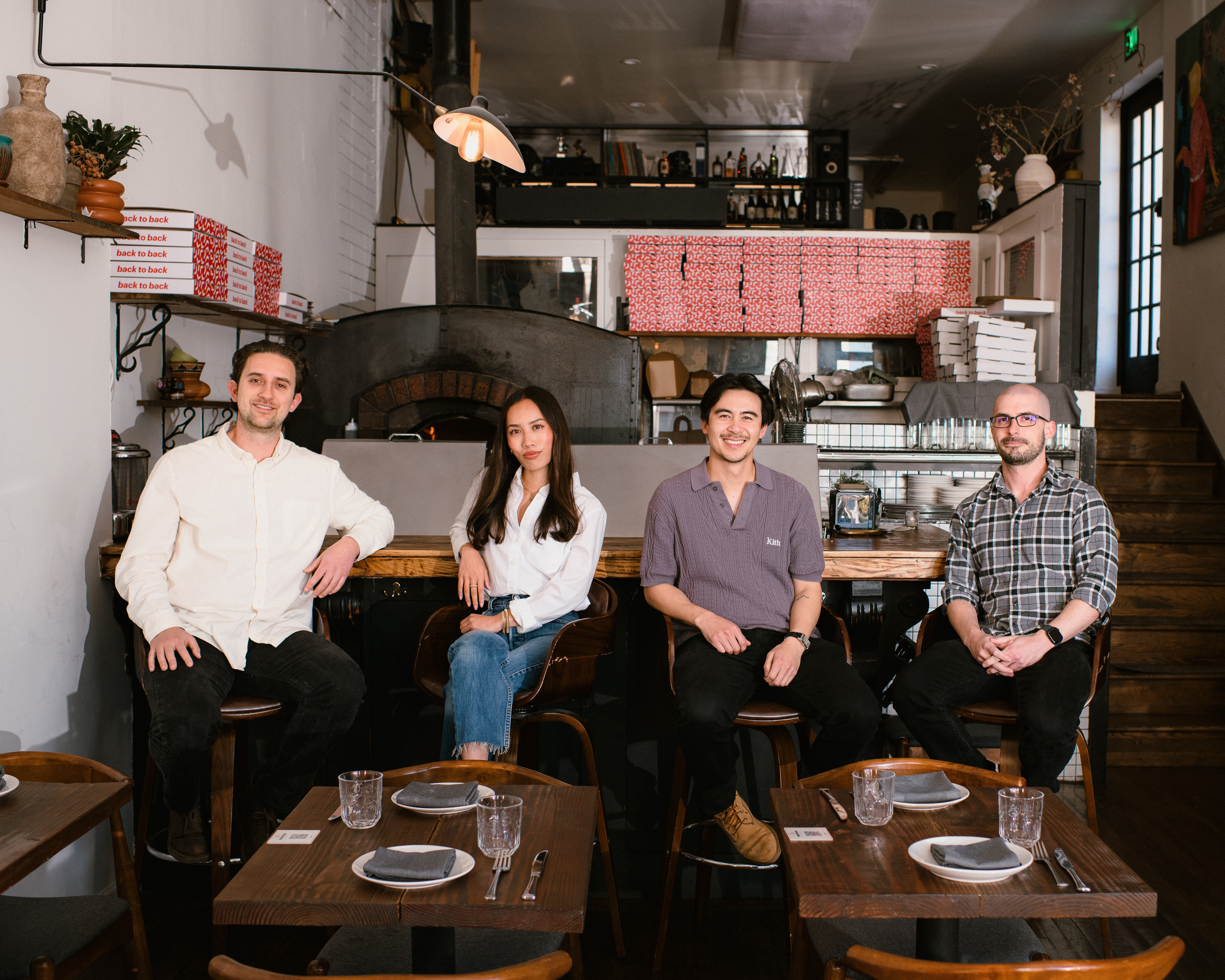 Four people are seated at a cozy restaurant counter. They smile towards the camera. The background includes stacked pizza boxes and a wood-fired oven.