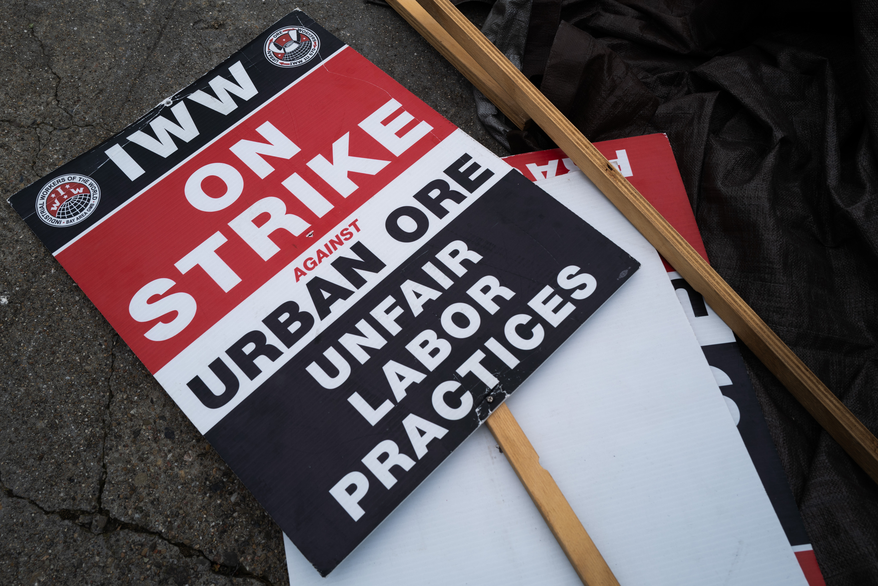 A protest sign on the ground reads &quot;IWW ON STRIKE against Urban Ore UNFAIR LABOR PRACTICES&quot; with bold red, black, and white colors.