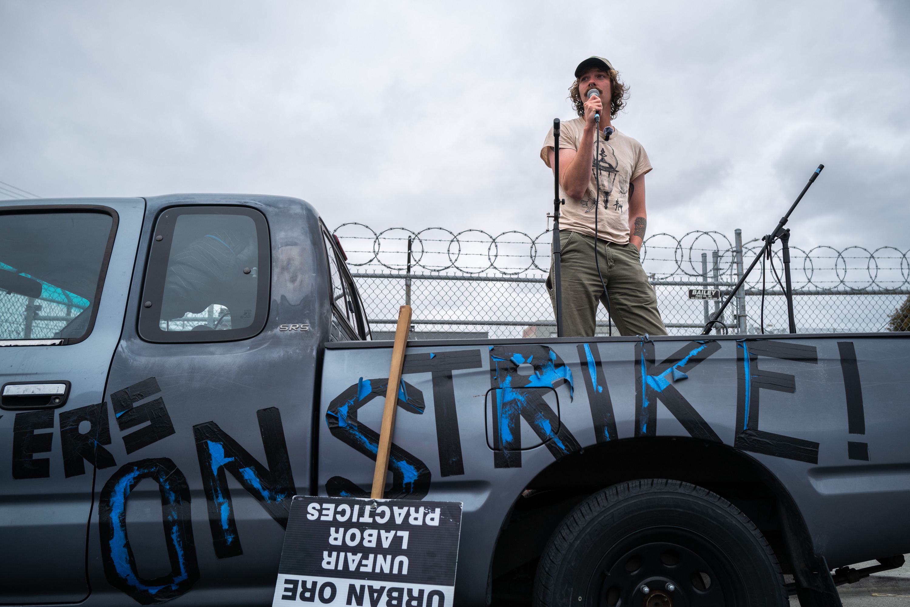 A person stands in a pickup truck bed holding a microphone, with &quot;ON STRIKE!&quot; painted on the truck. A sign about unfair labor practices leans against it.