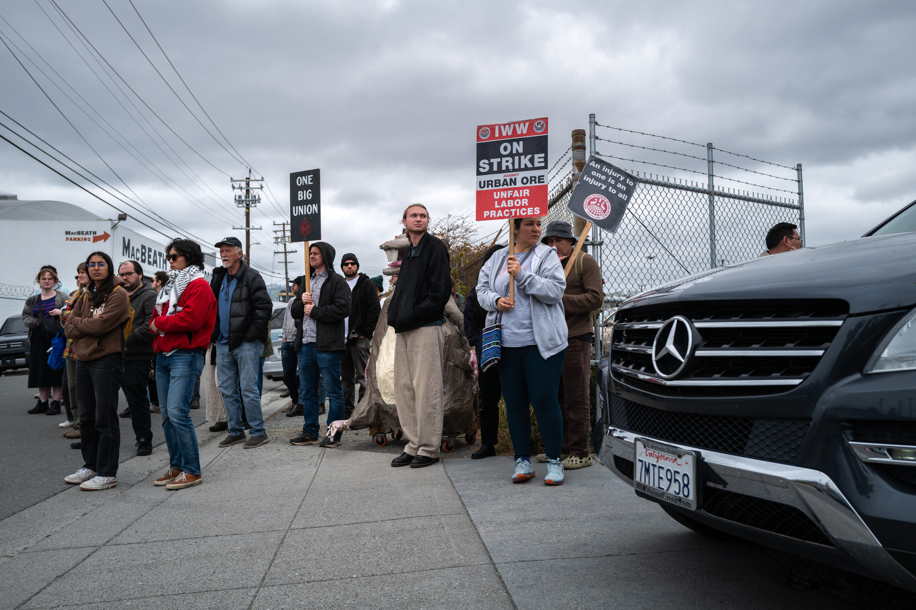 A group of people stand on a sidewalk holding signs about a labor strike, with a cloudy sky and a parked Mercedes nearby.