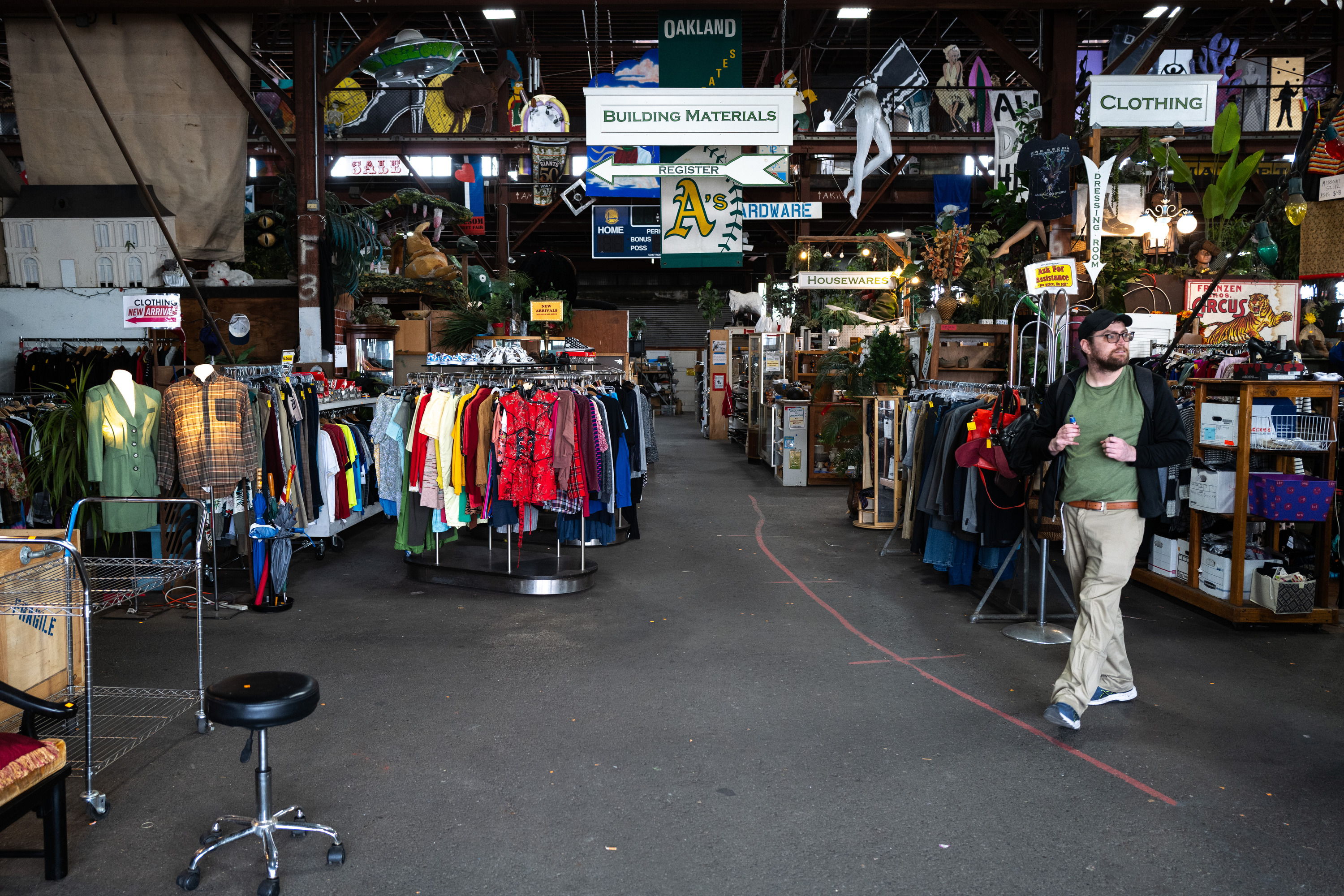 The image shows an indoor thrift store with racks of colorful clothing. Signs overhead indicate sections like clothing, housewares, and building materials. A person walks by.