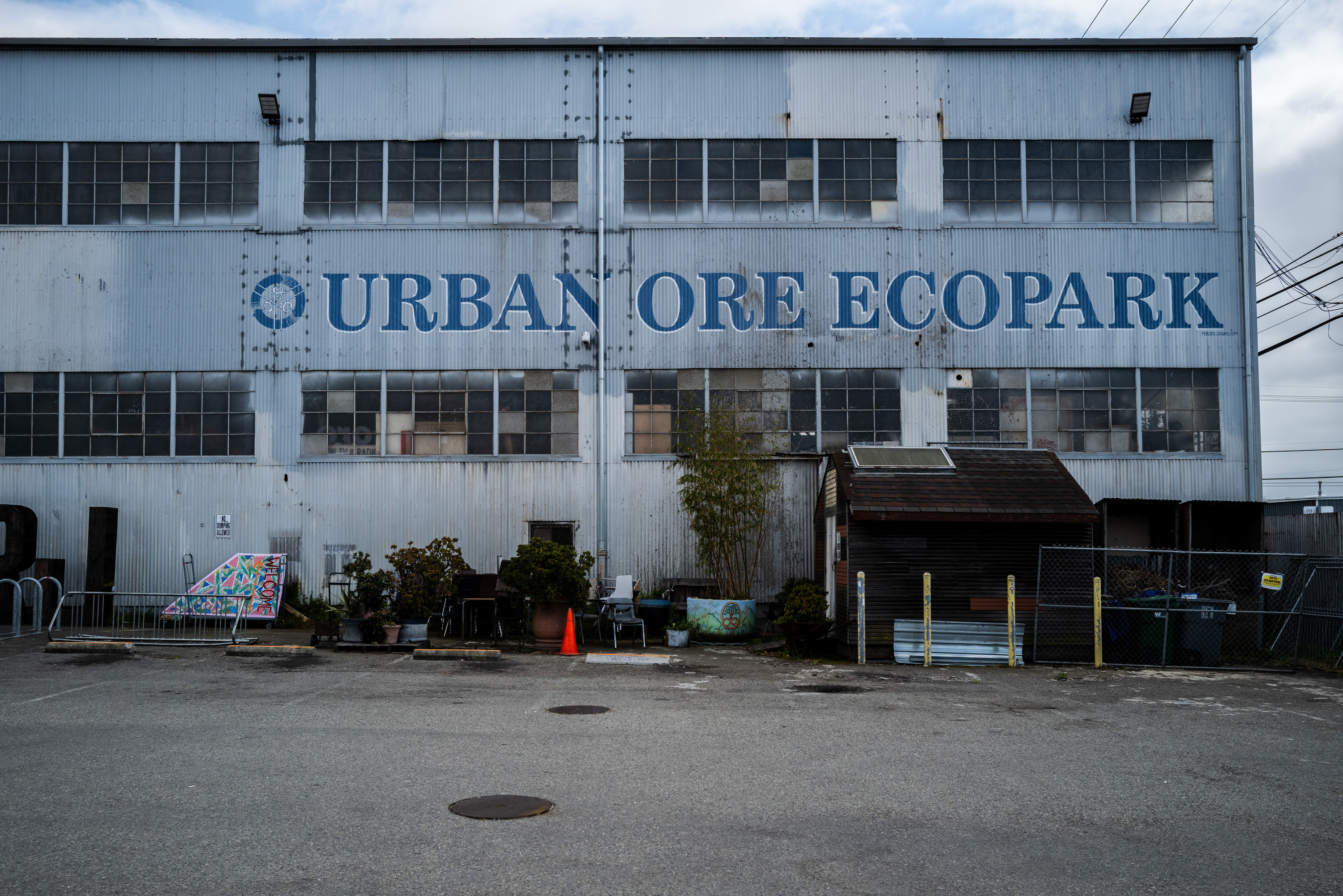 The image shows a large industrial building with &quot;Urban Ore Ecopark&quot; painted on it. The area has plants, a colorful sign, a wooden shed, and a traffic cone.
