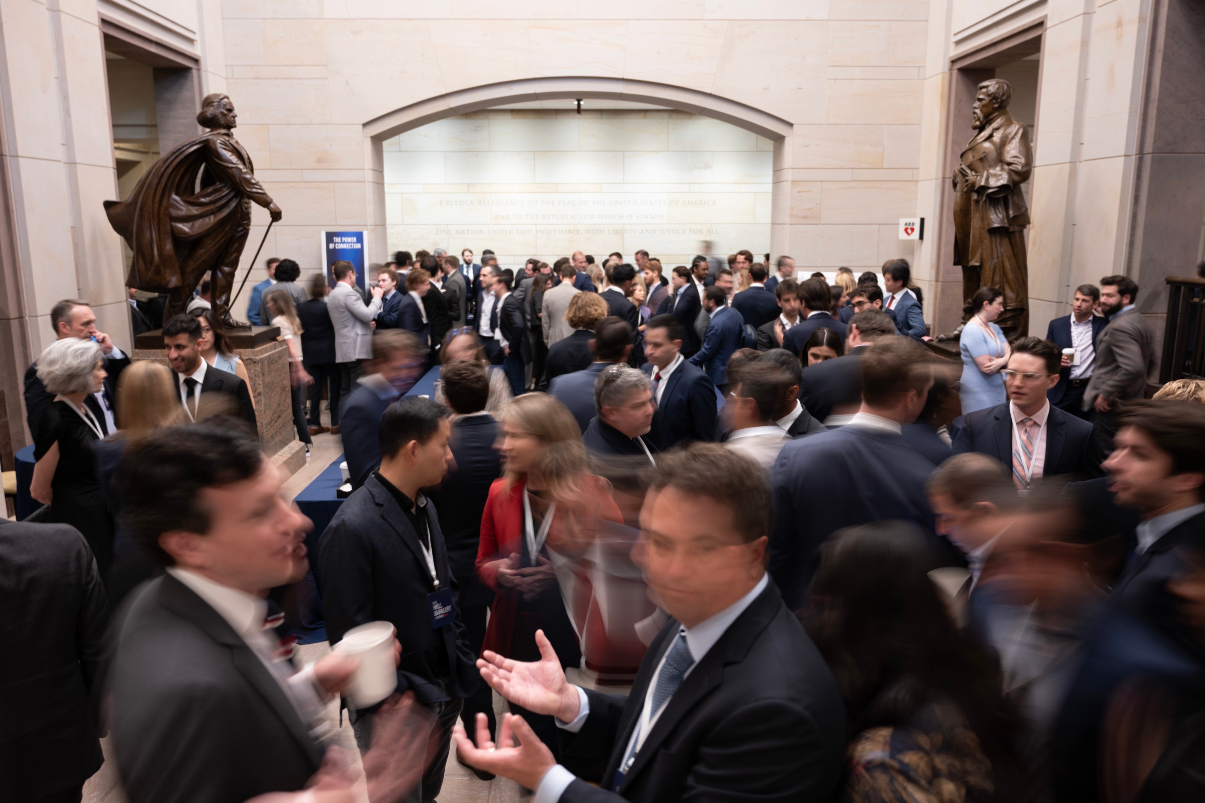 A crowded gathering takes place in a hall with statues, where people in formal attire are conversing and networking. The scene is lively and bustling.