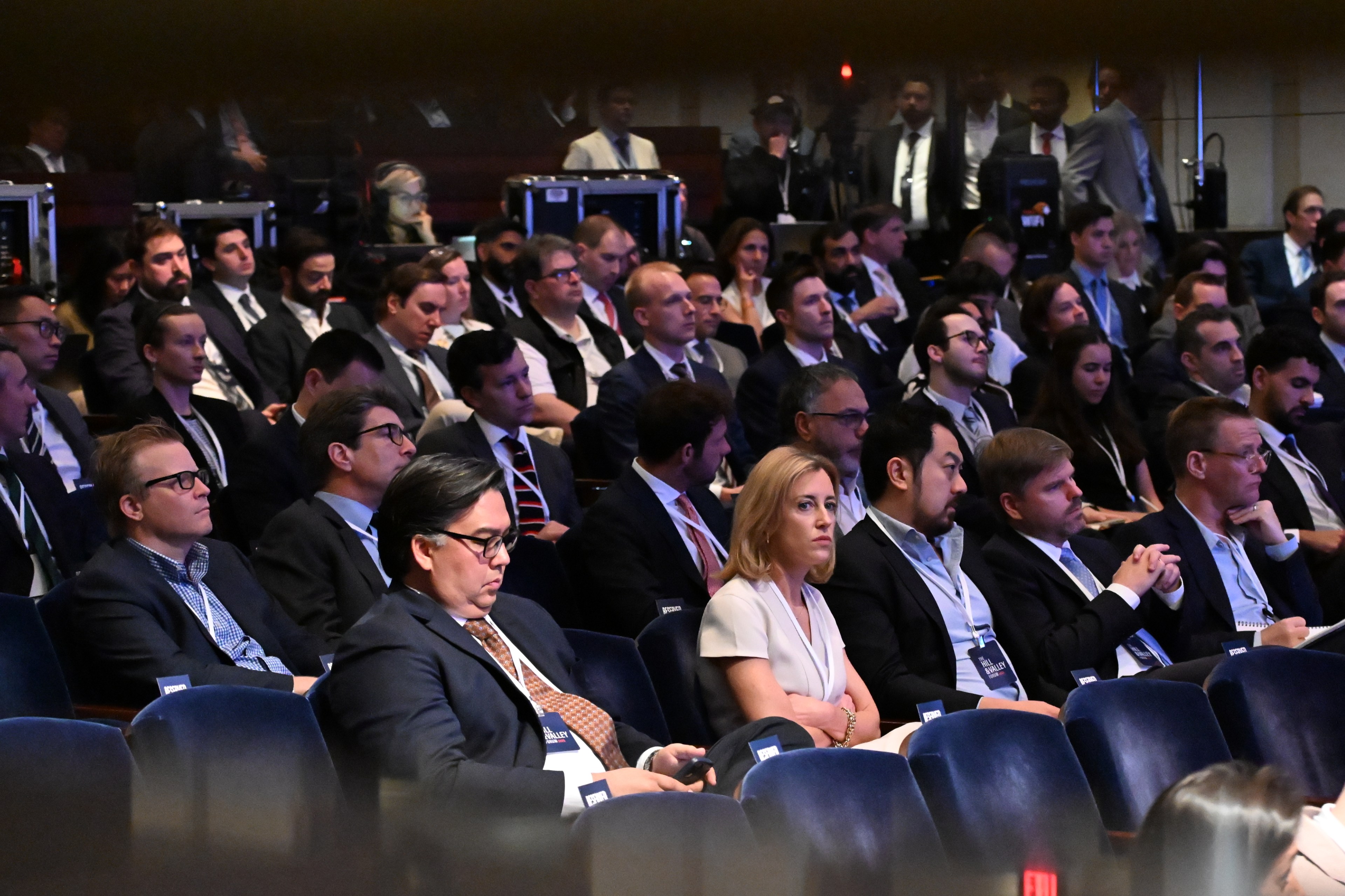 A large group of people, mostly in formal attire, sit in a dimly lit auditorium, attentively facing the stage. They wear conference badges around their necks.