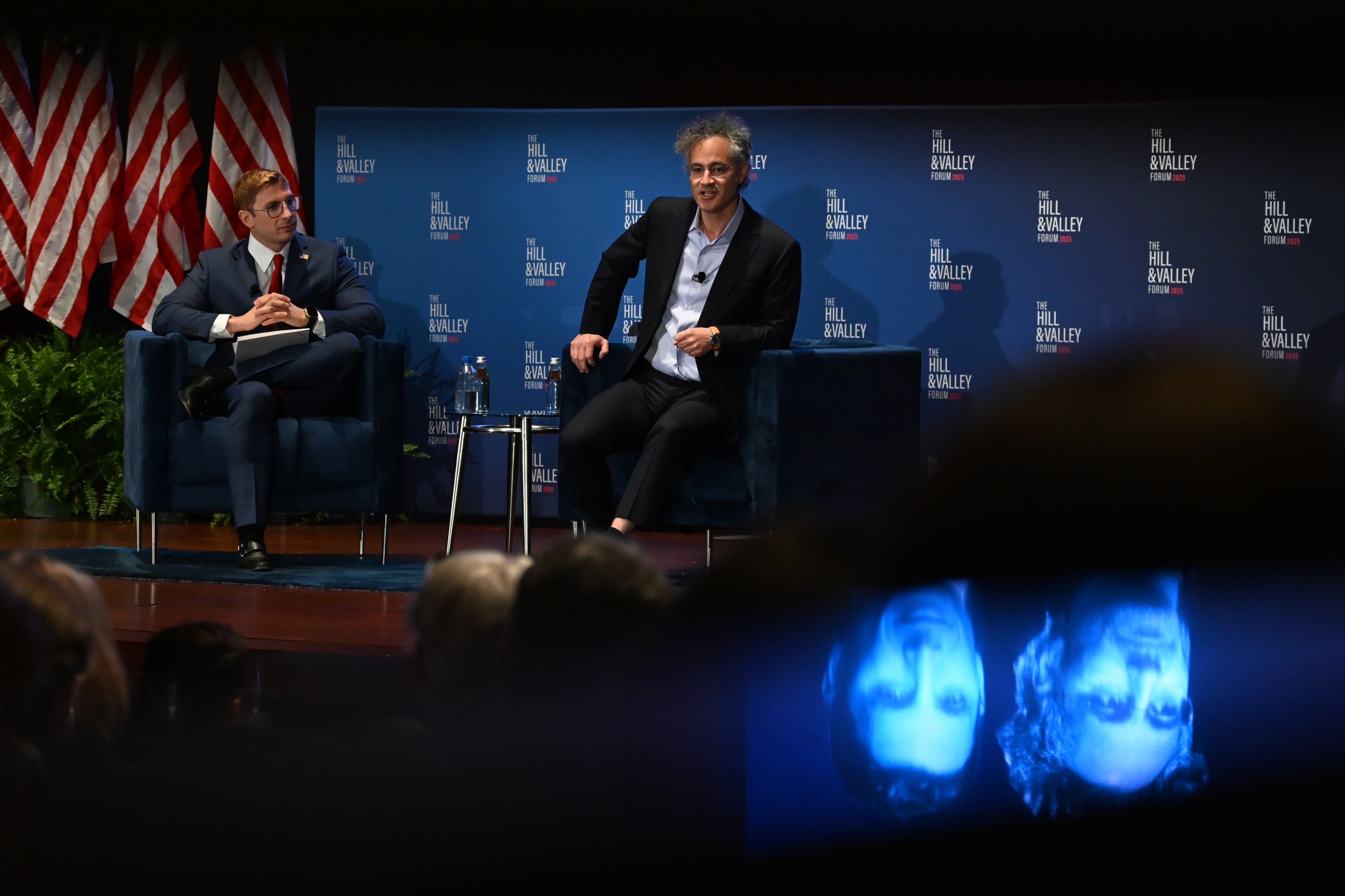 Two people are seated on a stage with &quot;The Hill &amp; Valley Forum&quot; backdrop. One wears a suit, and the other is casually dressed. U.S. flags line the background.