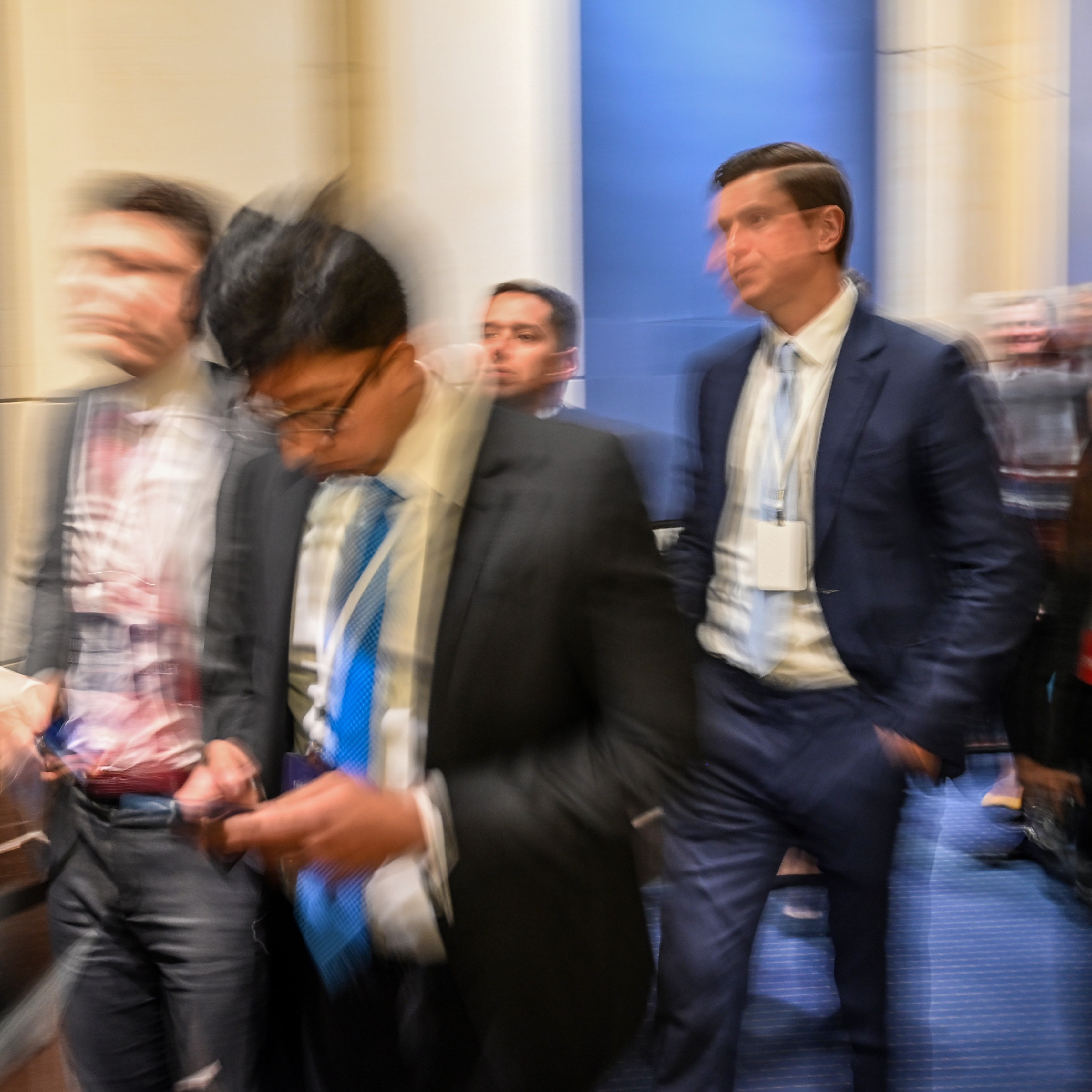A group of people in suits and business attire move quickly through a hallway, creating a blurred effect that suggests motion.