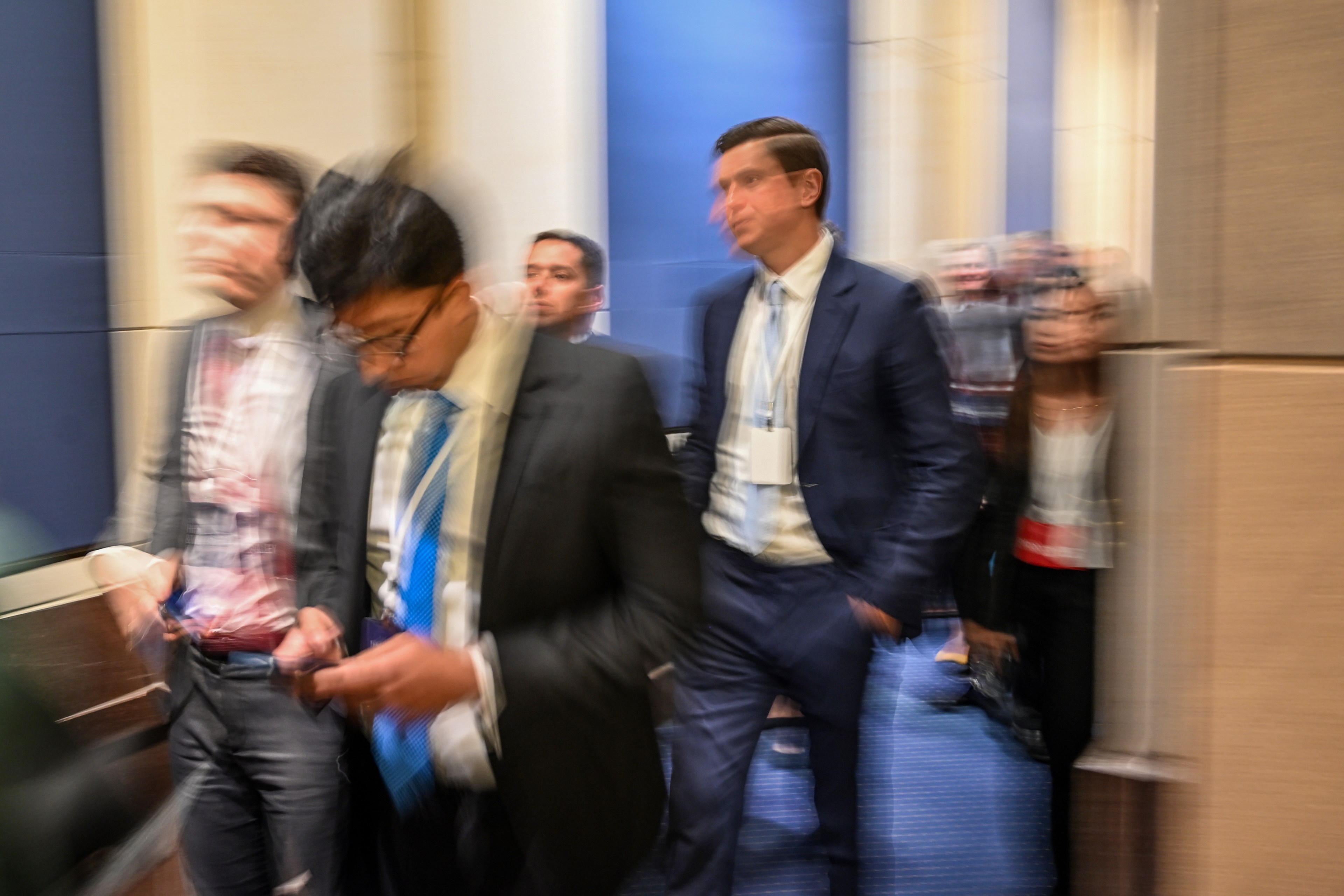 A group of people in suits and business attire move quickly through a hallway, creating a blurred effect that suggests motion.