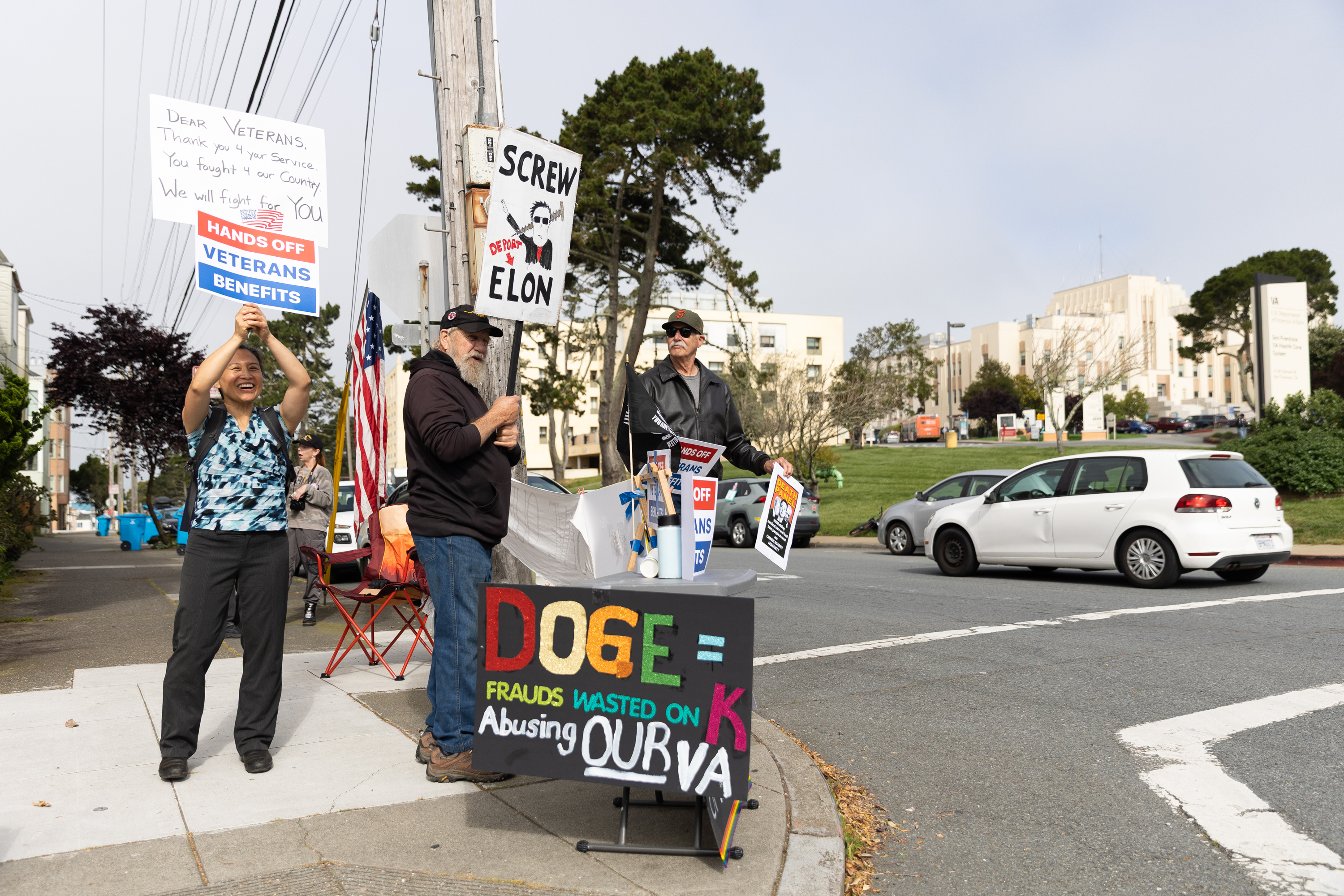Three protesters stand on a sidewalk holding signs criticizing Elon Musk and advocating for veterans' benefits, while a car passes by on the street.