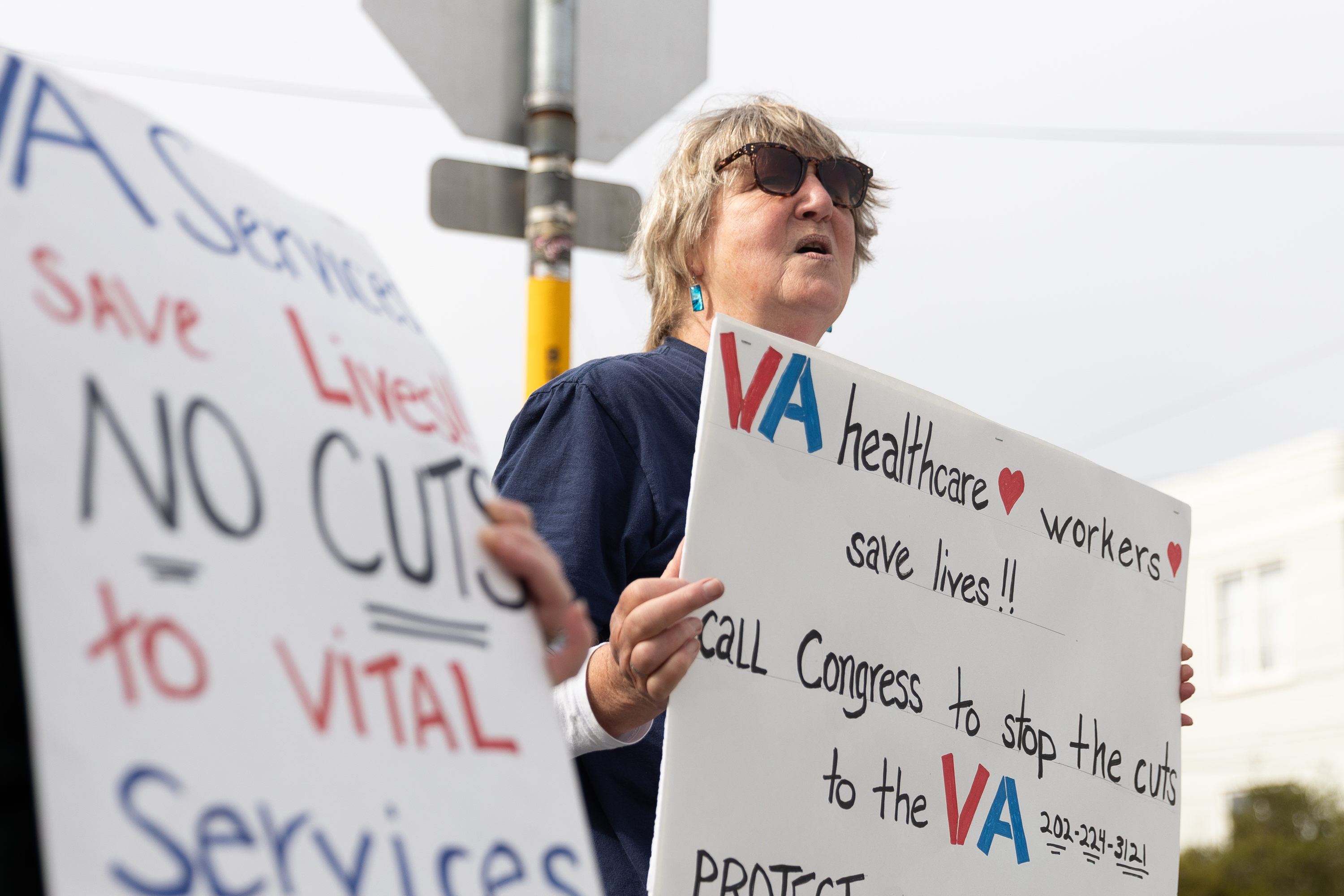 A person holds signs protesting cuts to VA healthcare services, emphasizing their importance in saving lives.