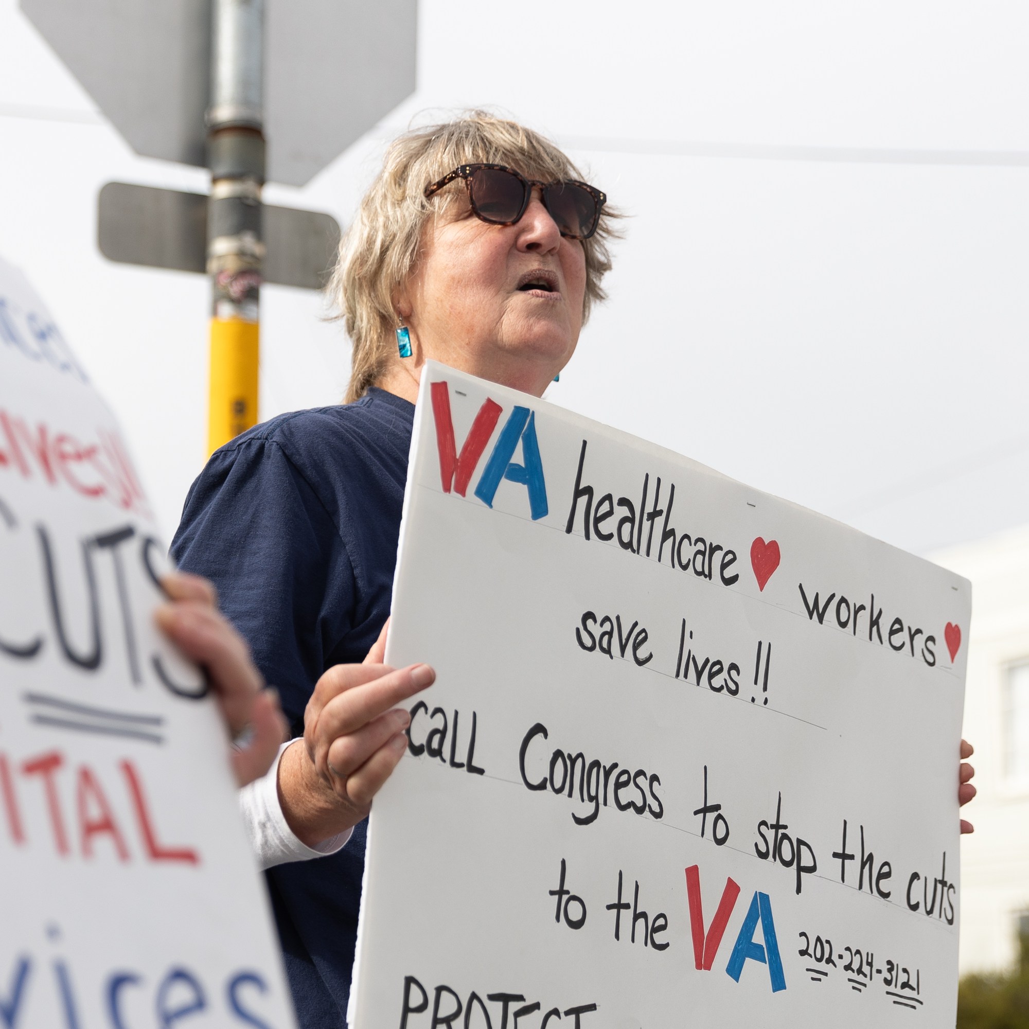 A person holds signs protesting cuts to VA healthcare services, emphasizing their importance in saving lives.