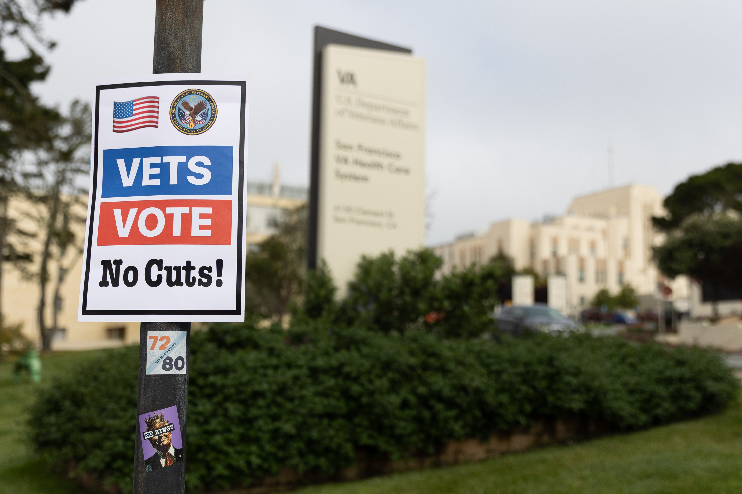 A sign with American flags and &quot;Vets Vote No Cuts!&quot; is on a pole, with a building and trees in the blurred background.