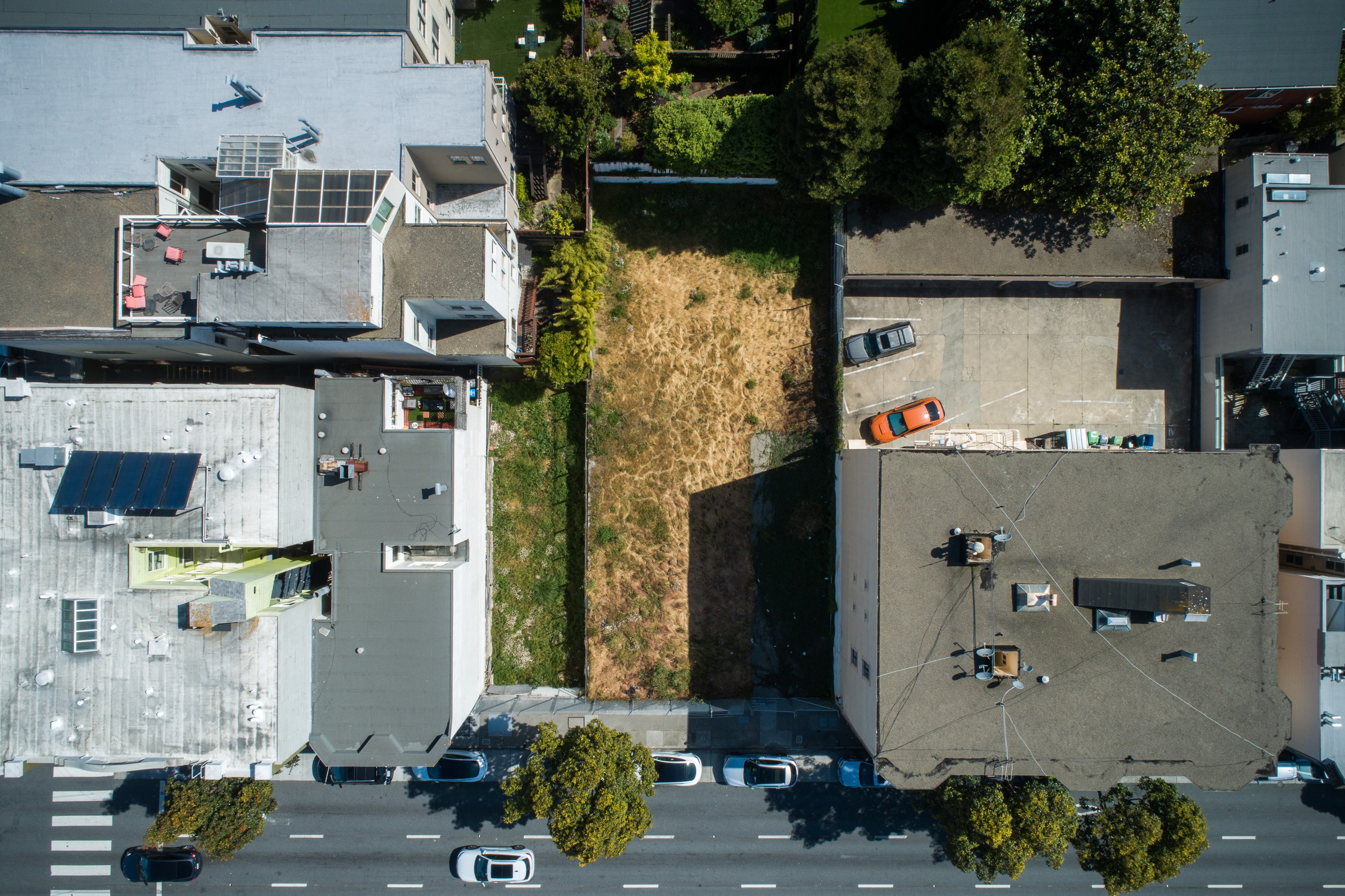 The image shows a bird's-eye view of an urban area, featuring two buildings with flat roofs, a dry grassy lot between them, and cars parked on the street.