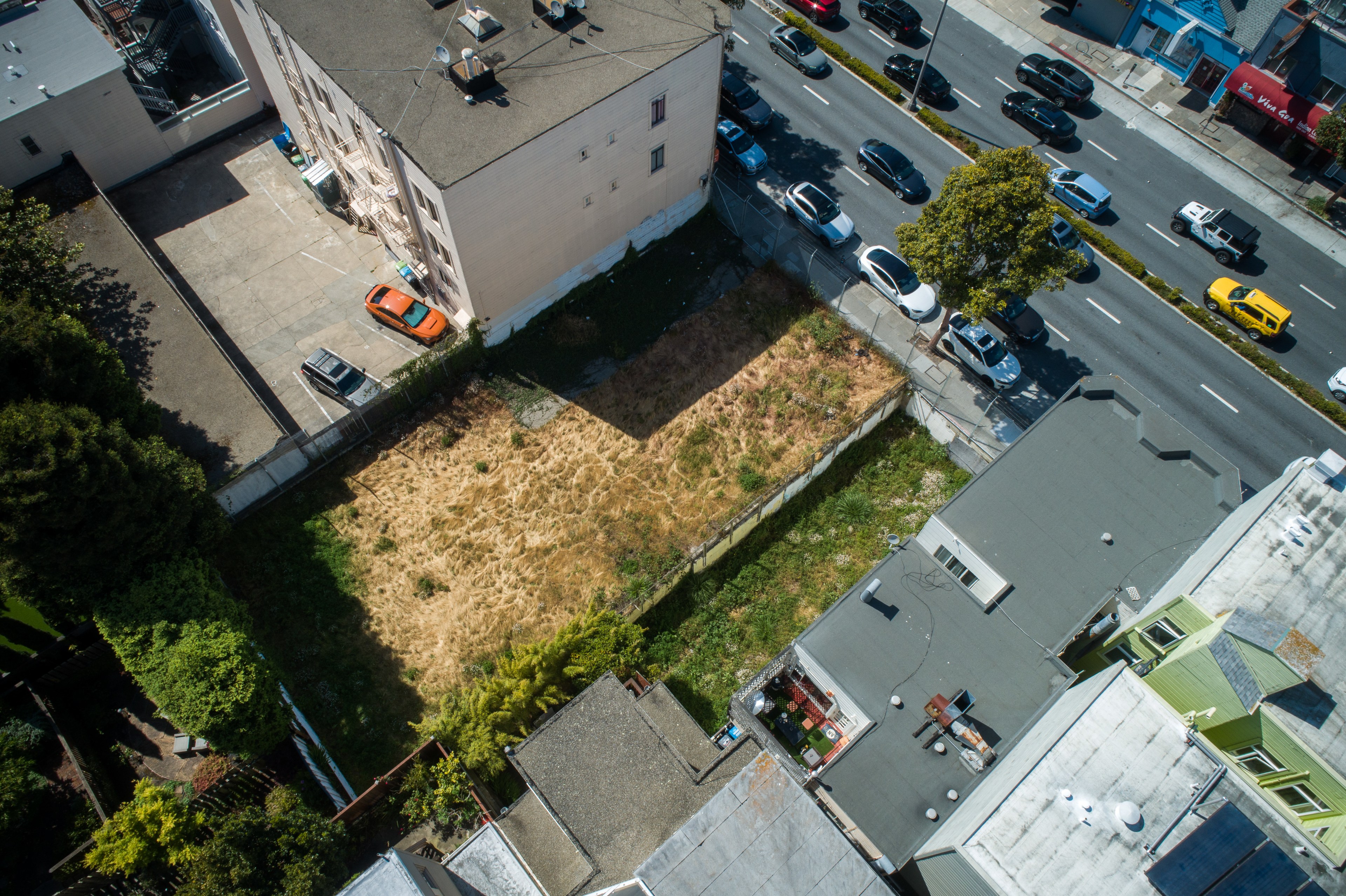 The image shows an aerial view of an empty, overgrown lot surrounded by buildings and a street with parked cars and traffic.