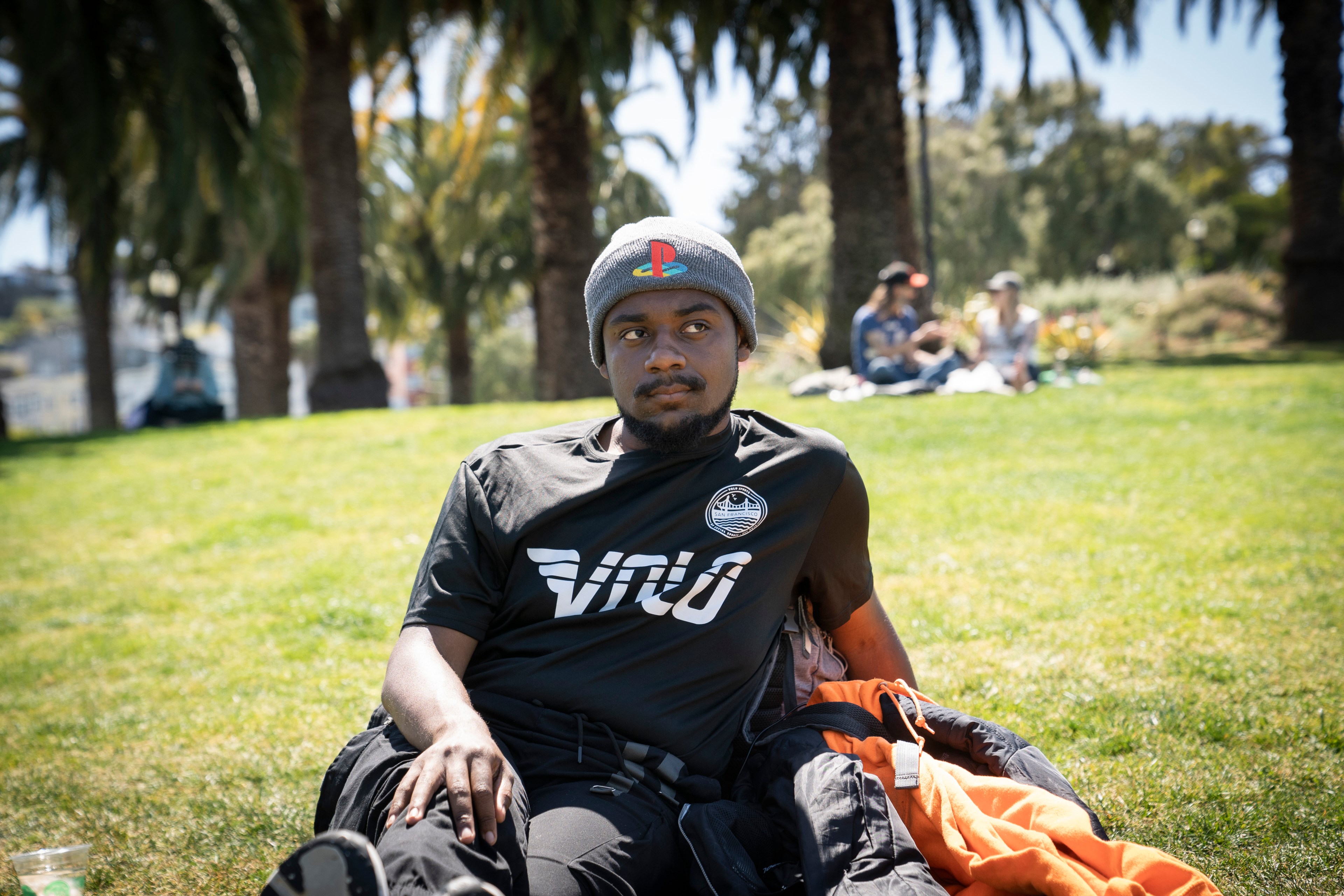 A person sits on grass in a park, wearing a gray beanie and black shirt with logos. Palm trees are in the background, and people relax on the grass.