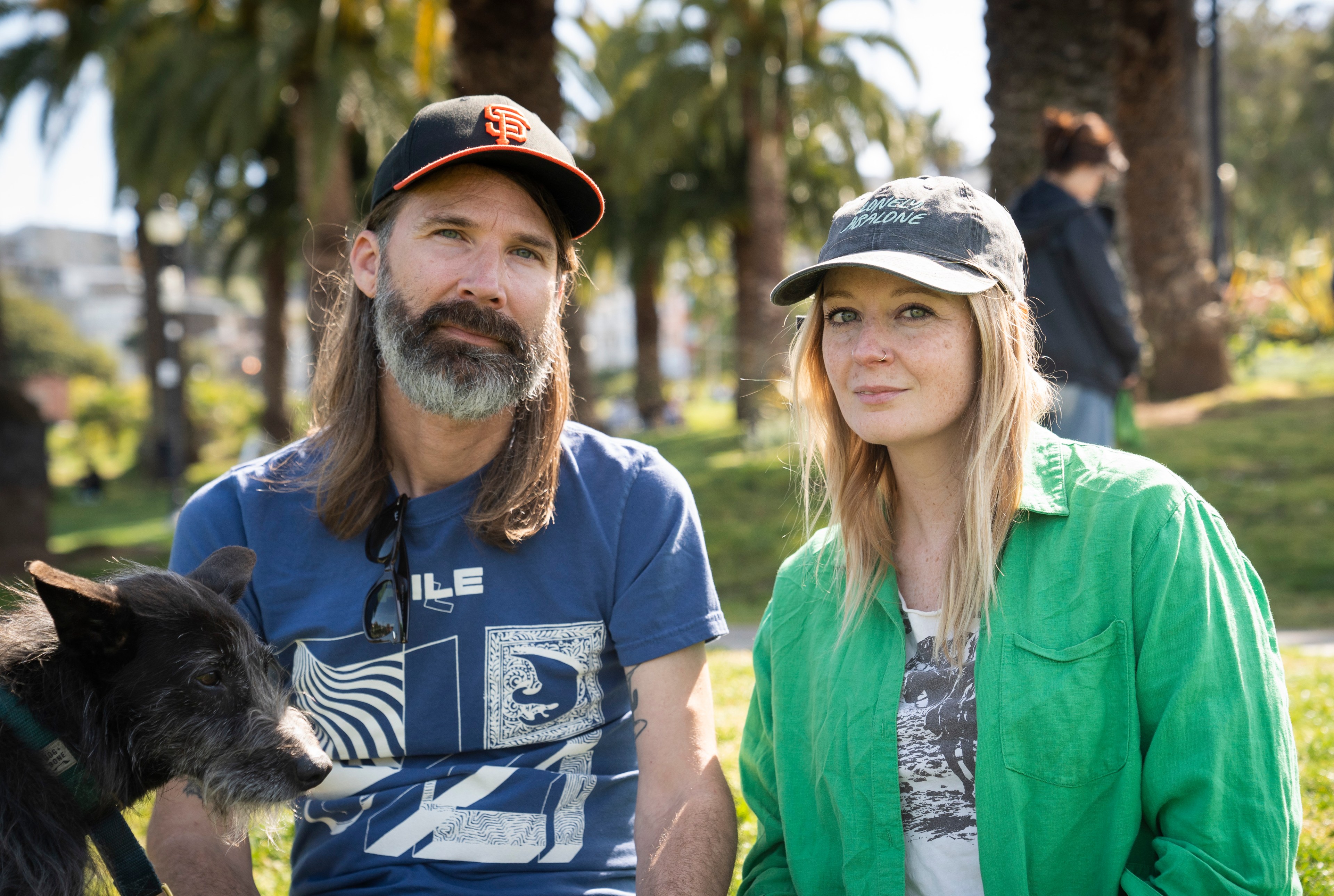 A man with a beard and a cap and a woman with freckles in a green shirt, both sitting outside with a black dog nearby. Palm trees are in the background.