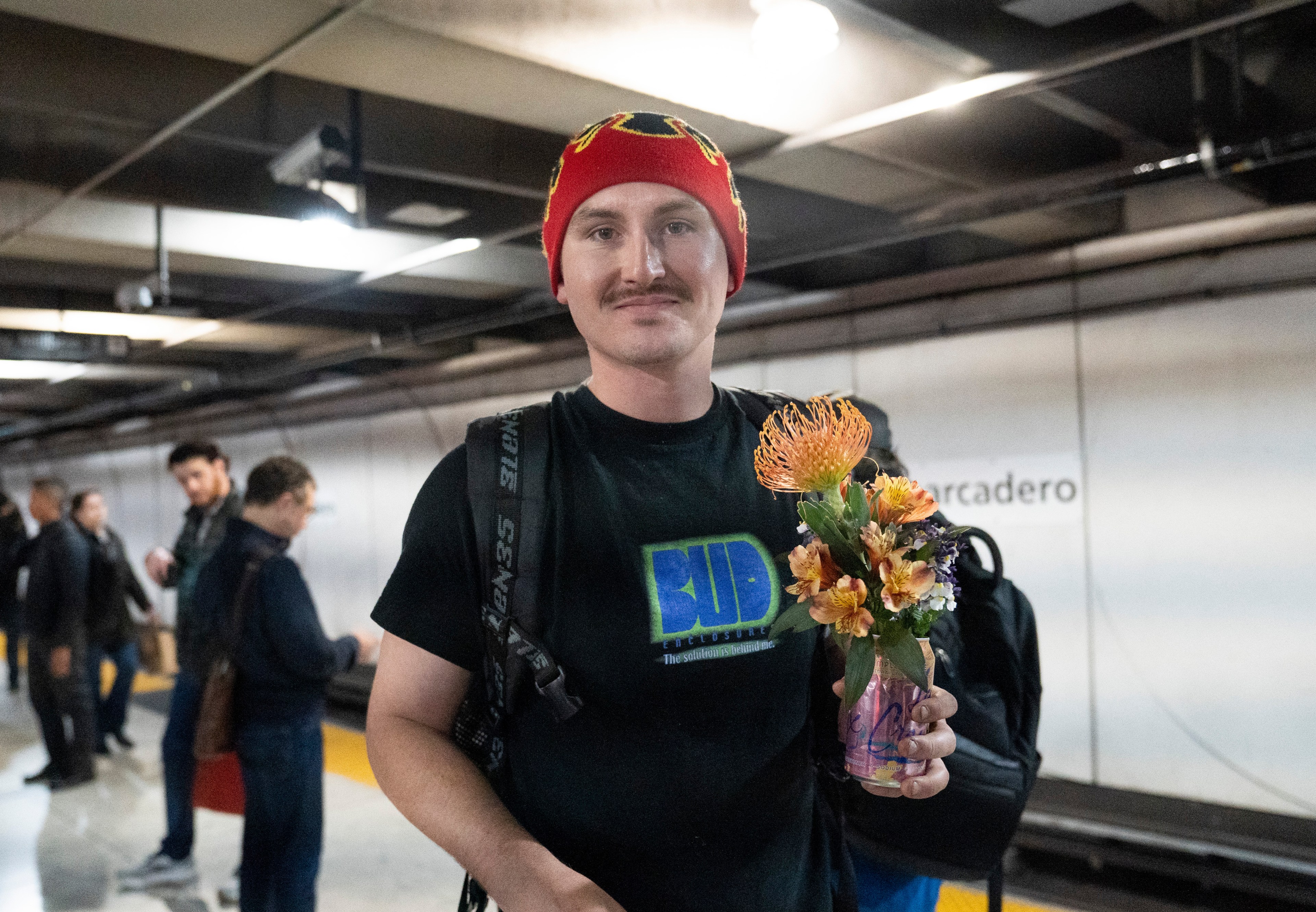 A person in a red beanie and black t-shirt holds a colorful bouquet in a can, standing on a subway platform with others in the background.