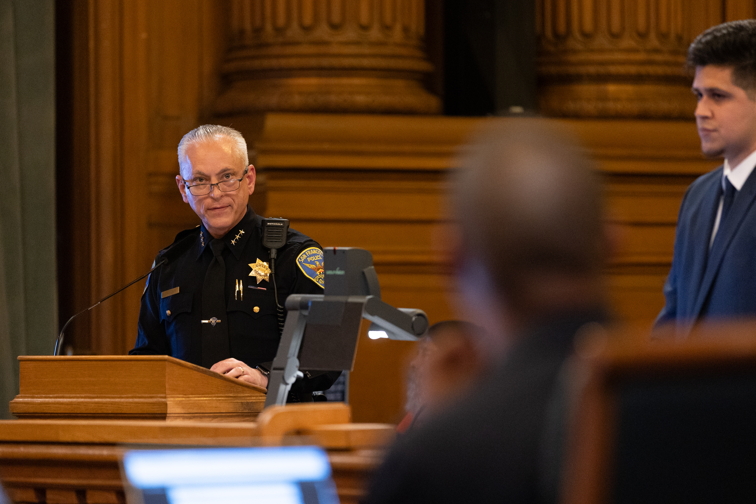 A police officer in uniform speaks at a wooden podium with a badge visible. A man in a suit stands to the right. The setting appears formal, likely a courtroom.