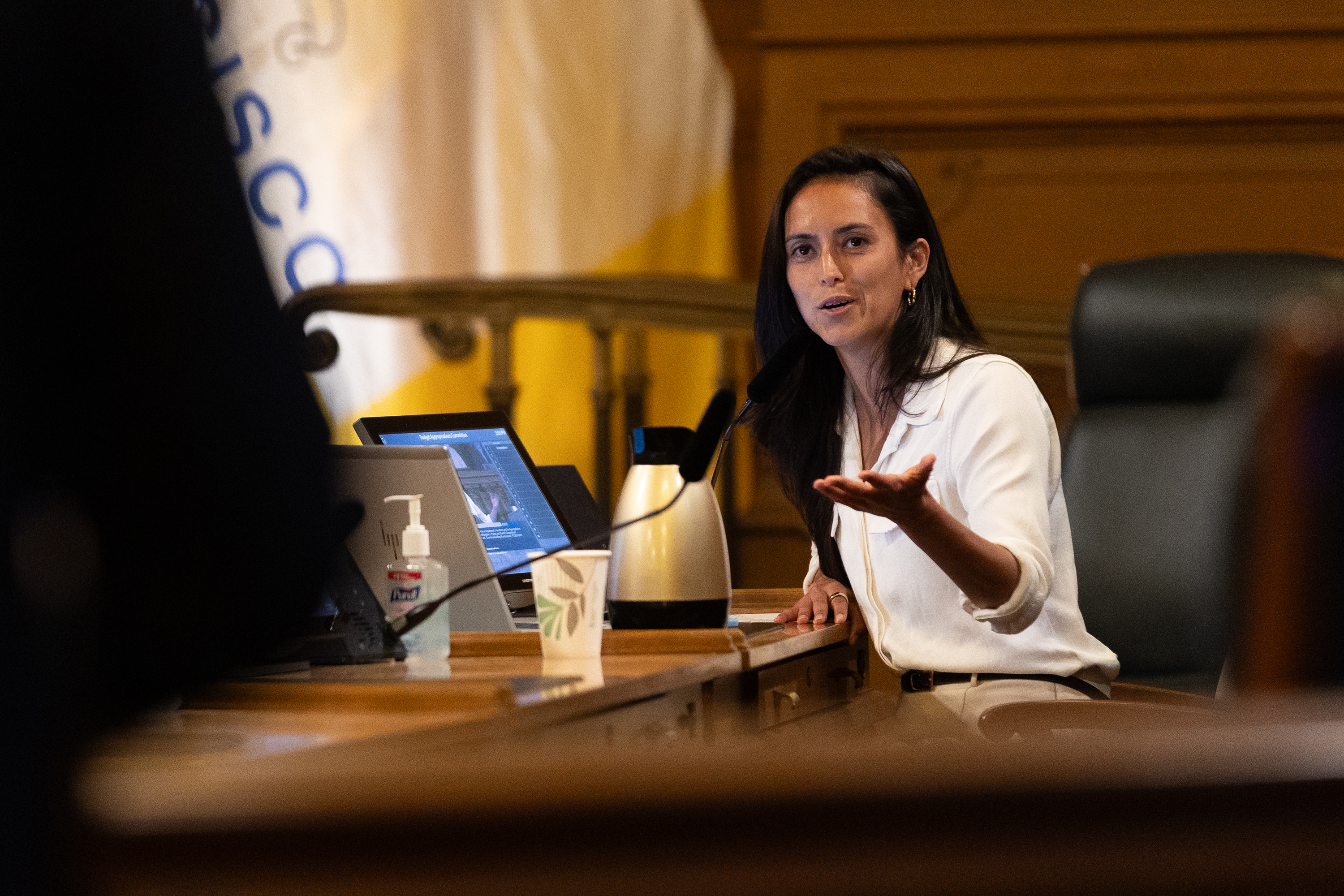 A woman sits at a table with a laptop, a coffee carafe, and hand sanitizer, speaking into a microphone. She gestures with one hand, looking engaged.