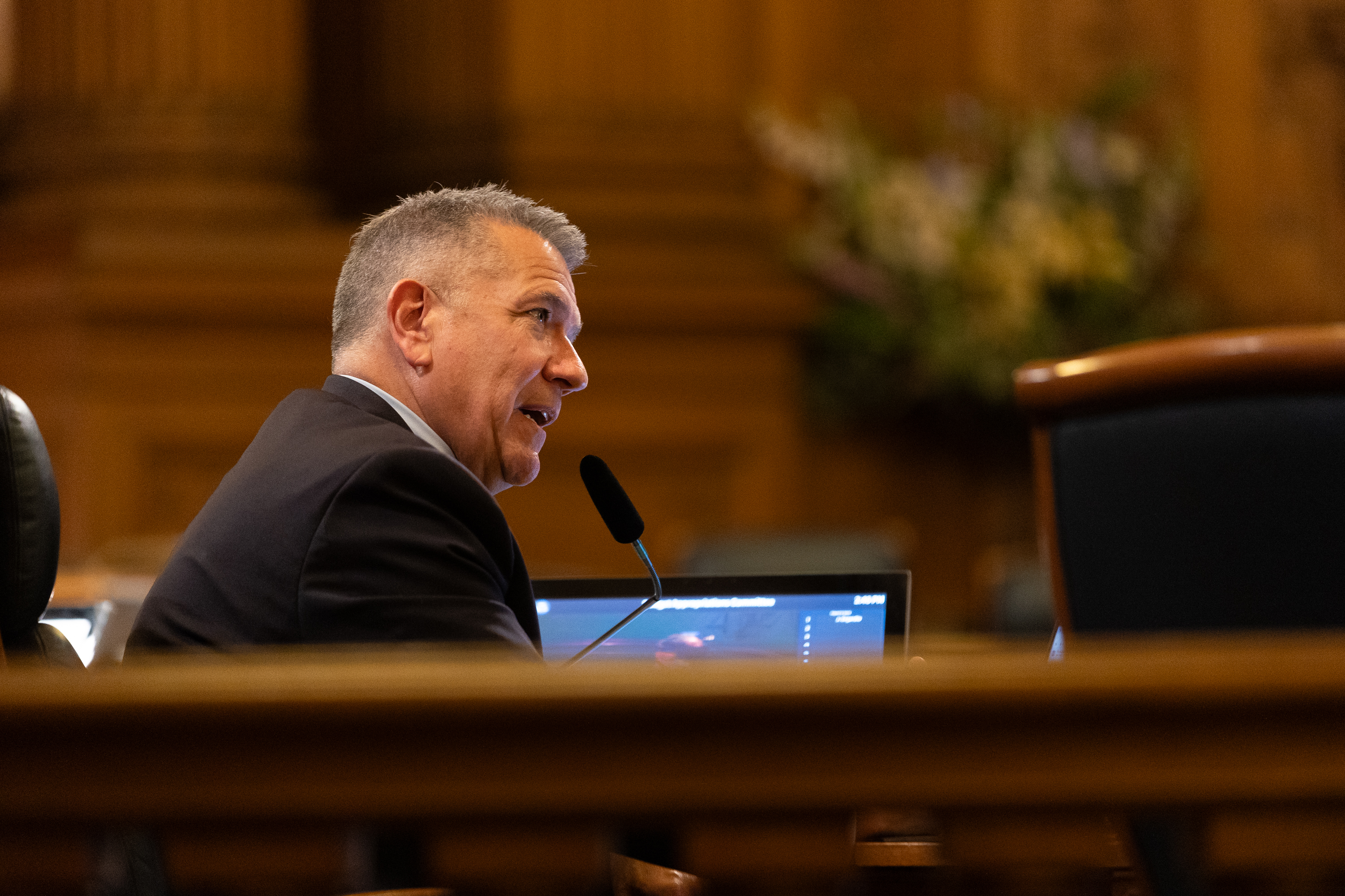 A person in a suit is speaking into a microphone in a formal setting with wood-paneled walls and a computer screen in front of them.