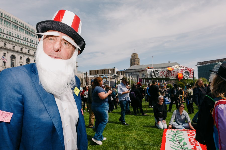 A person dressed as Uncle Sam stands in a park with a crowd. They wear a blue suit, white beard, and striped hat. Banners and people are visible in the background.