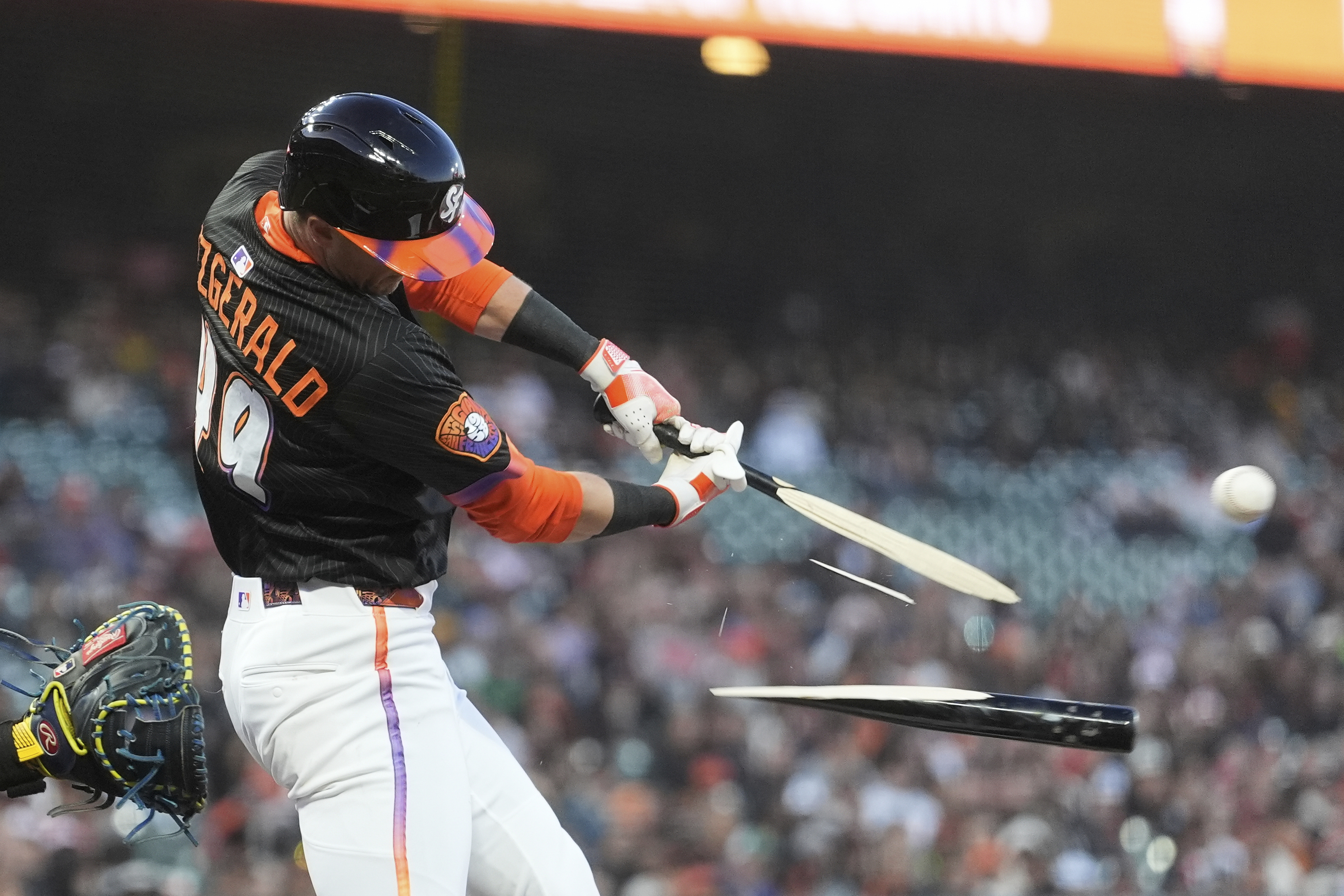 A baseball player in a black jersey swings and breaks his bat as he hits the ball, with the shattered bat pieces flying in opposite directions.