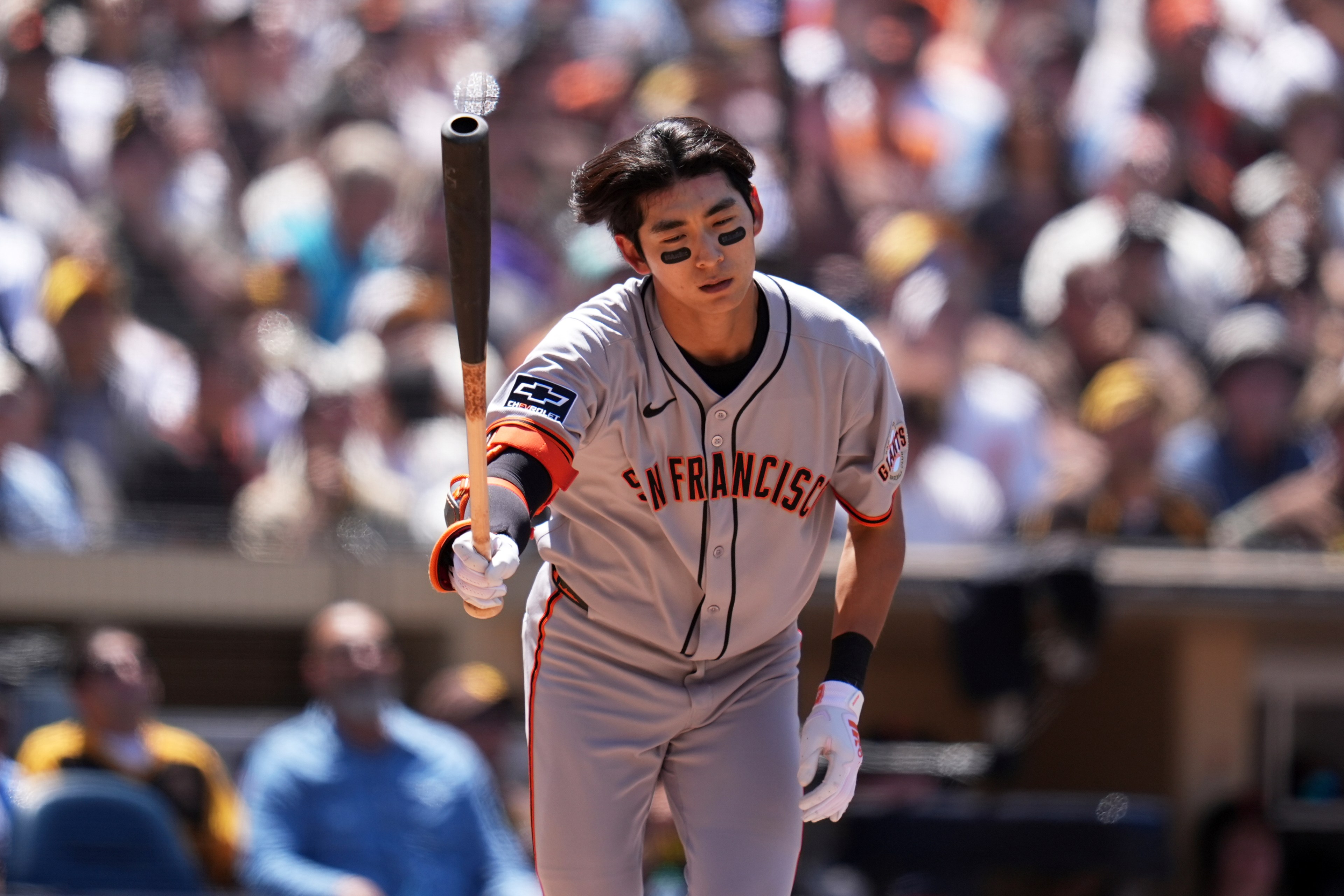 A baseball player in a gray uniform with "San Francisco" written on it holds a bat, with a focused expression. A blurred crowd is in the background.