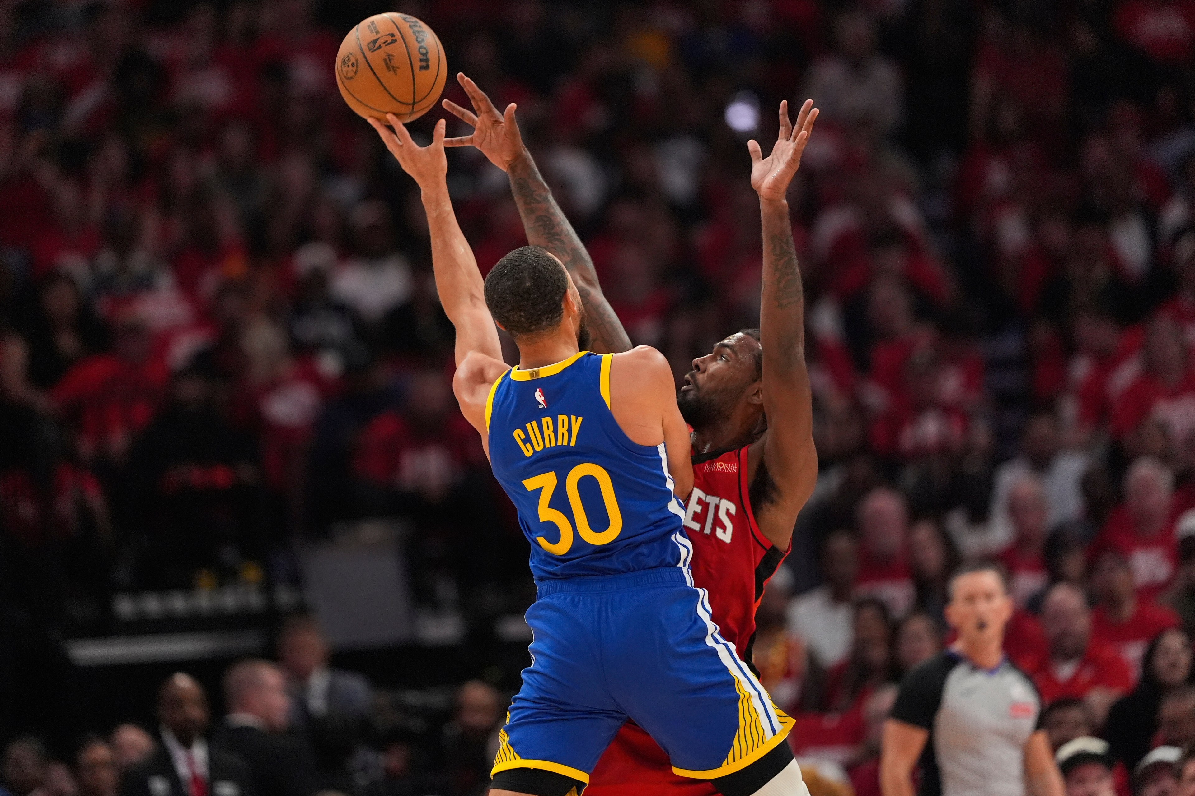 A basketball player in a blue jersey jumps to shoot, while another in a red jersey defends with arms raised. The crowd watches intensely from the background.