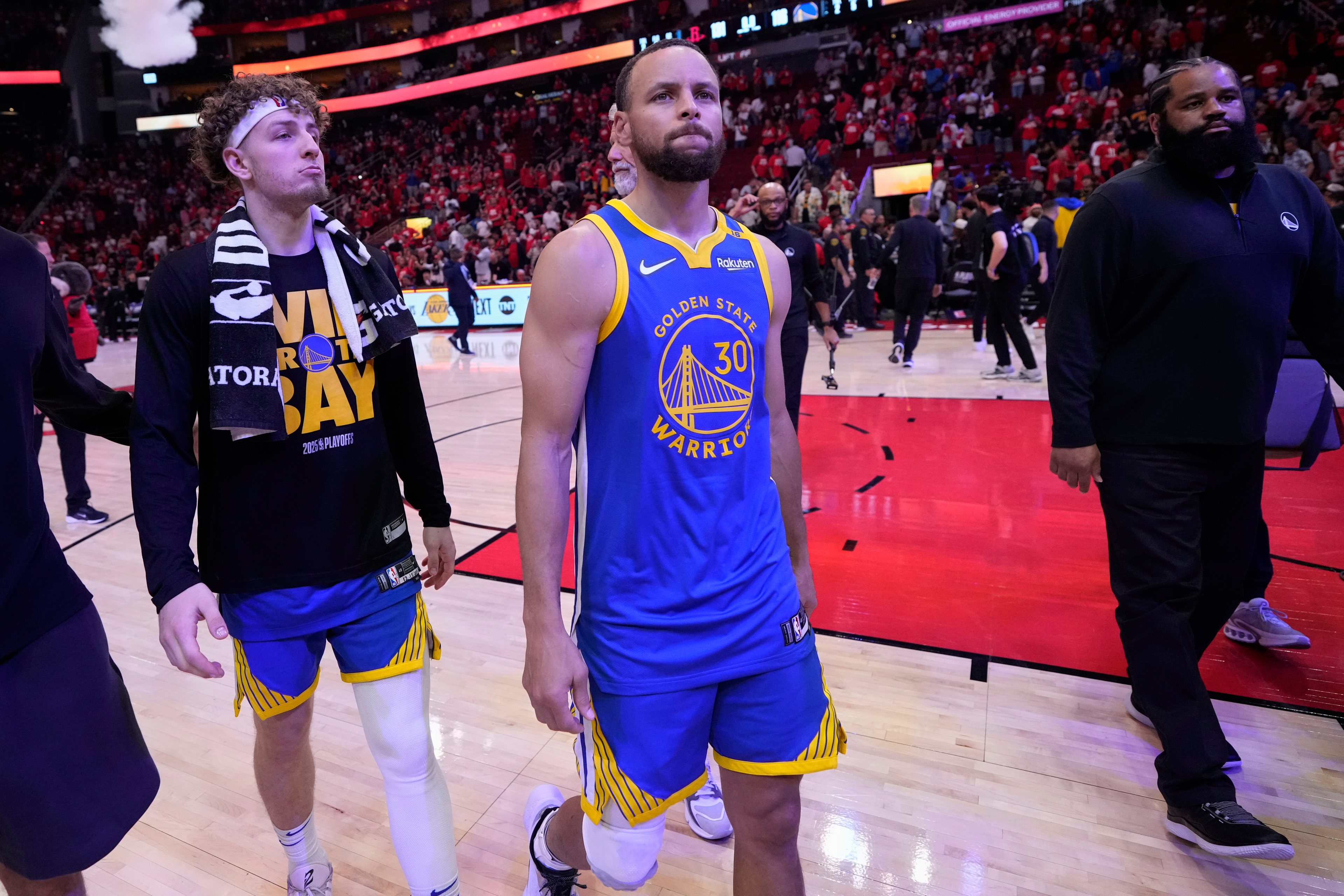 Two basketball players walk on the court, one in a blue Golden State Warriors jersey, the other in a team shirt with a towel over his shoulder, surrounded by a crowd.