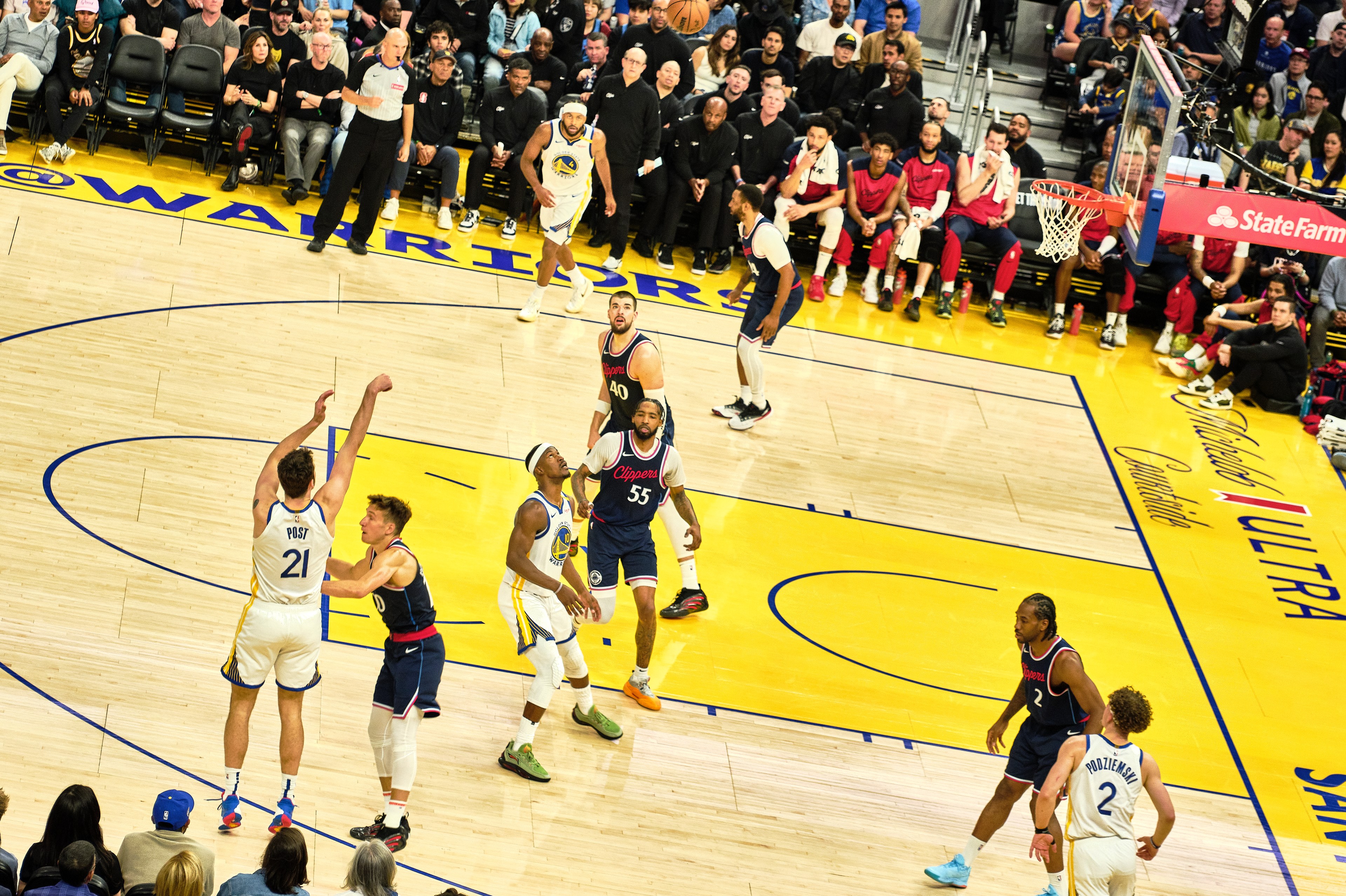 A basketball player in a white jersey takes a jump shot while others watch. The court is vibrant with a yellow and blue design, and the stands are full of spectators.