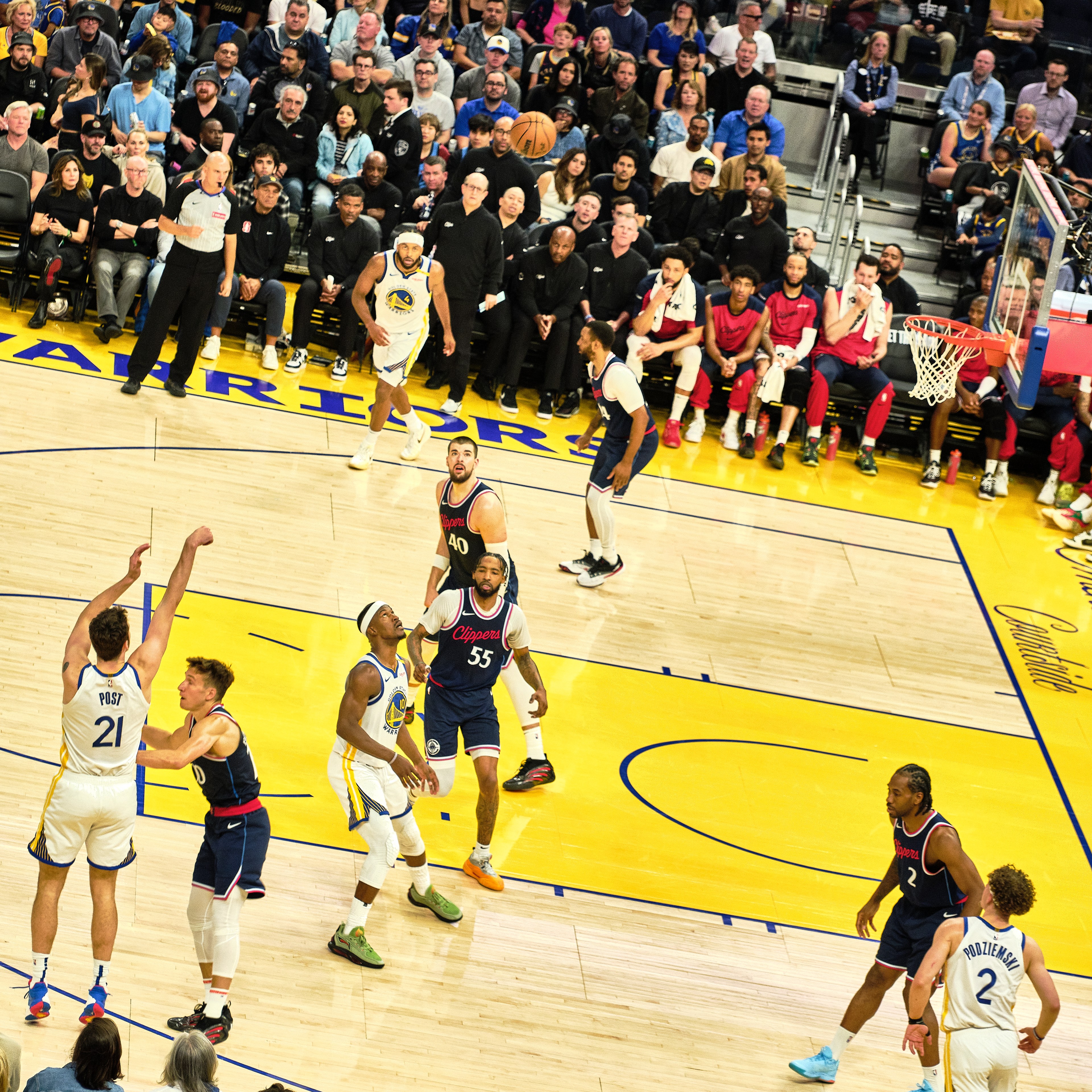 A basketball player in a white jersey takes a jump shot while others watch. The court is vibrant with a yellow and blue design, and the stands are full of spectators.