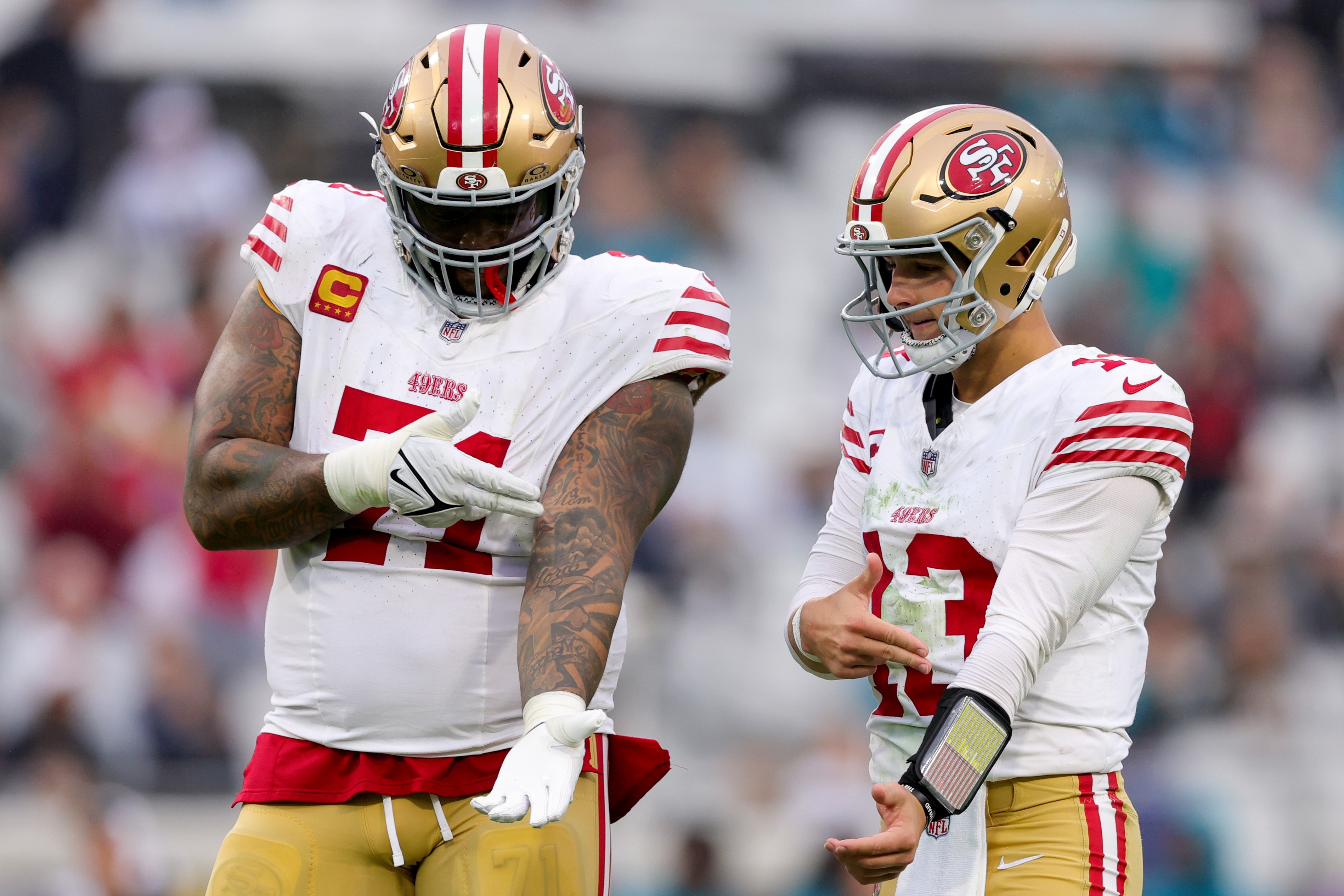 Two football players in white and red 49ers uniforms stand jokingly gesturing at each other on the field, wearing helmets and looking relaxed.