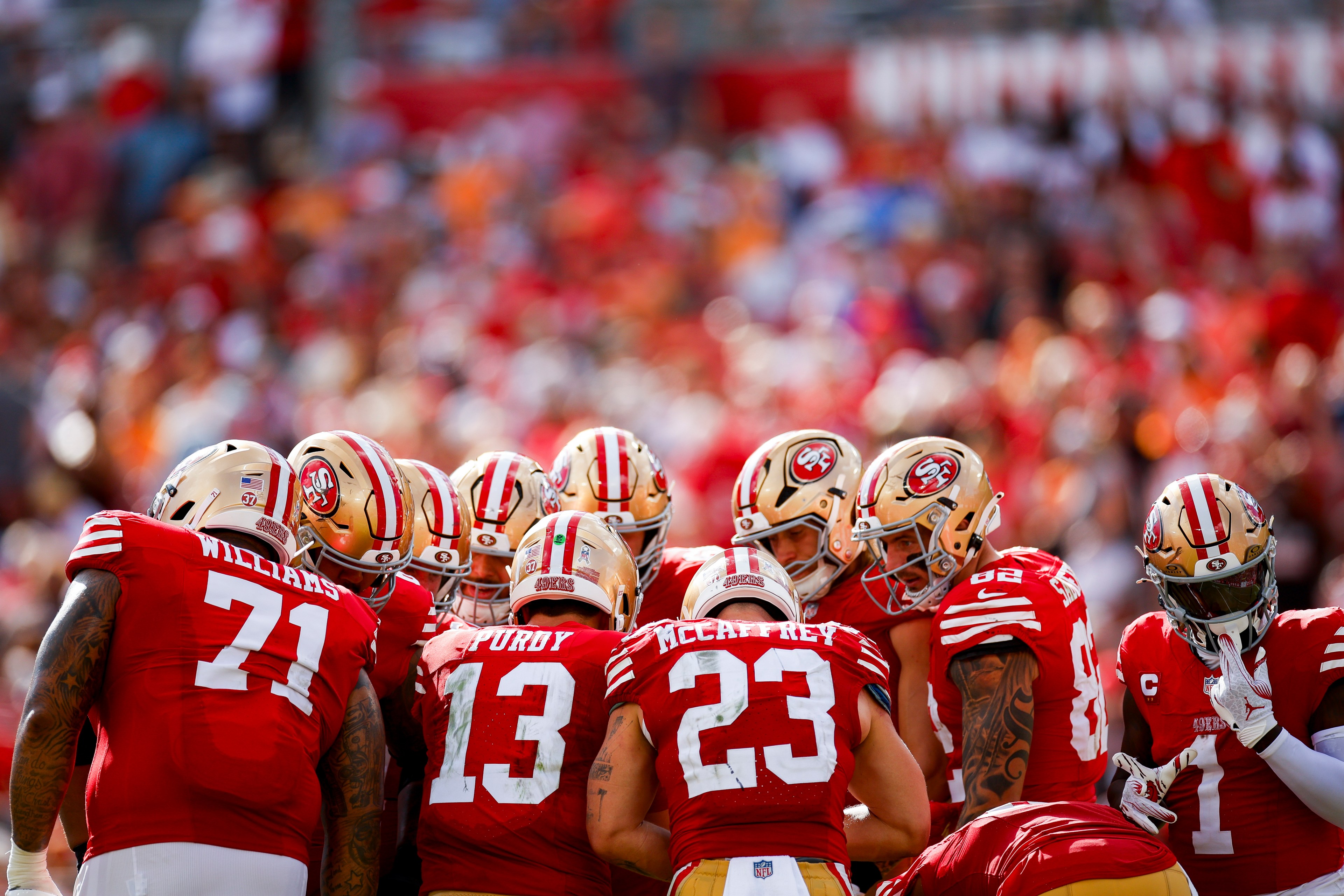 A group of football players in red and gold uniforms are huddled together on the field, focusing intently. Their jerseys display the numbers and names clearly.