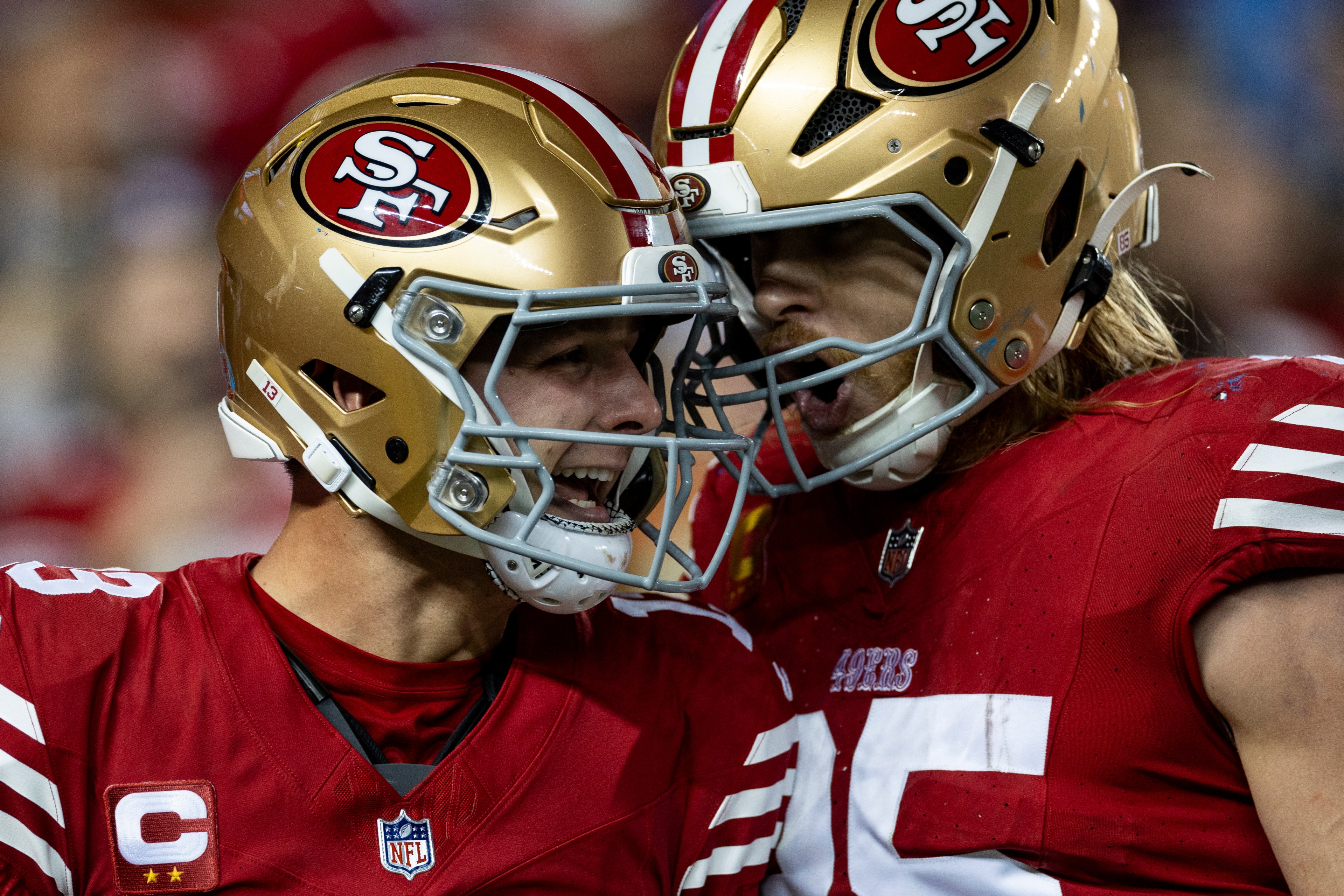 Two football players in red jerseys and gold helmets are celebrating energetically on the field. They are facing each other with intense expressions.