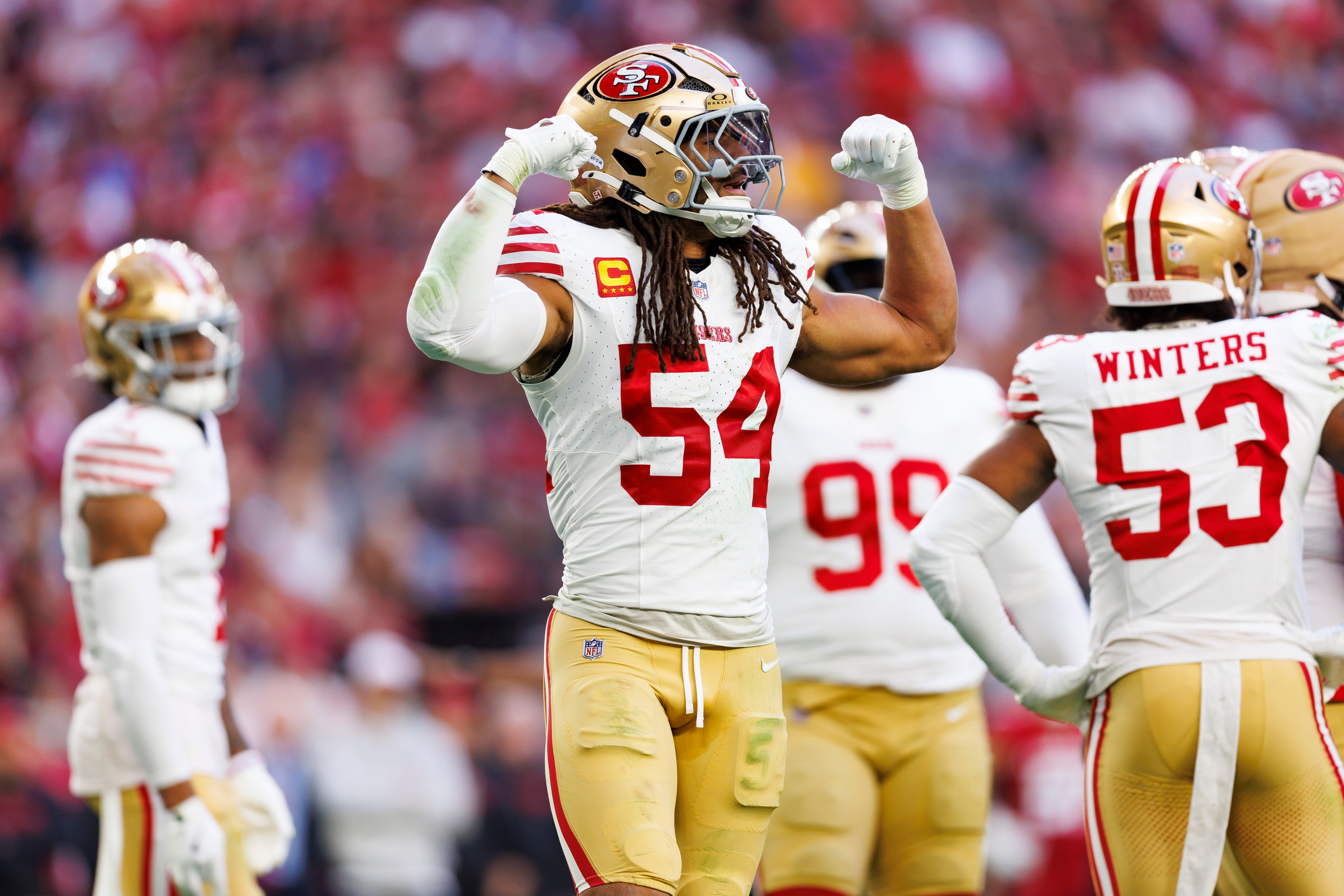 A football player in a white and red uniform flexes his muscles on the field, surrounded by teammates wearing similar gear, with a blurred crowd in the background.