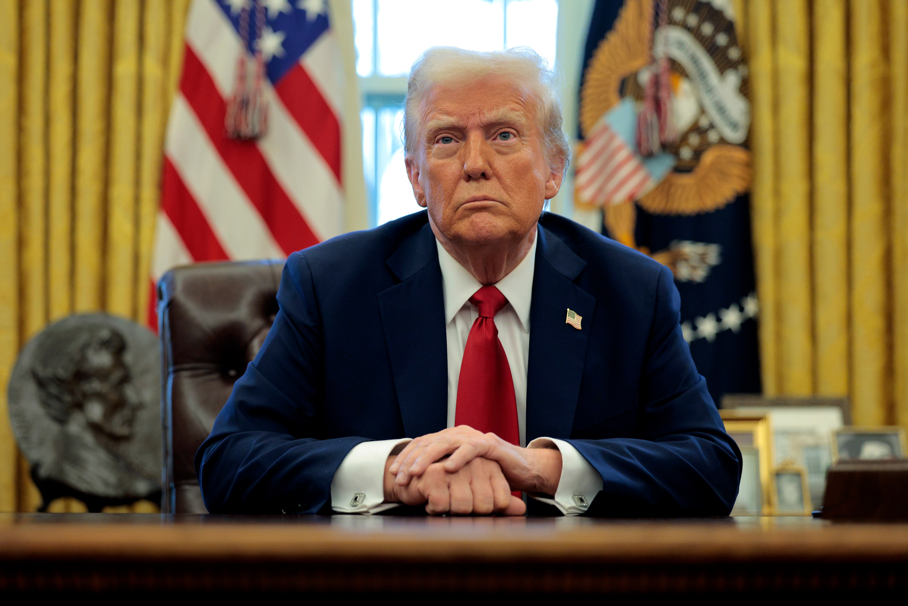 A man in a suit and red tie sits at a desk with hands clasped. Behind are American flags and a seal on gold curtains. A bust and framed photos are visible.