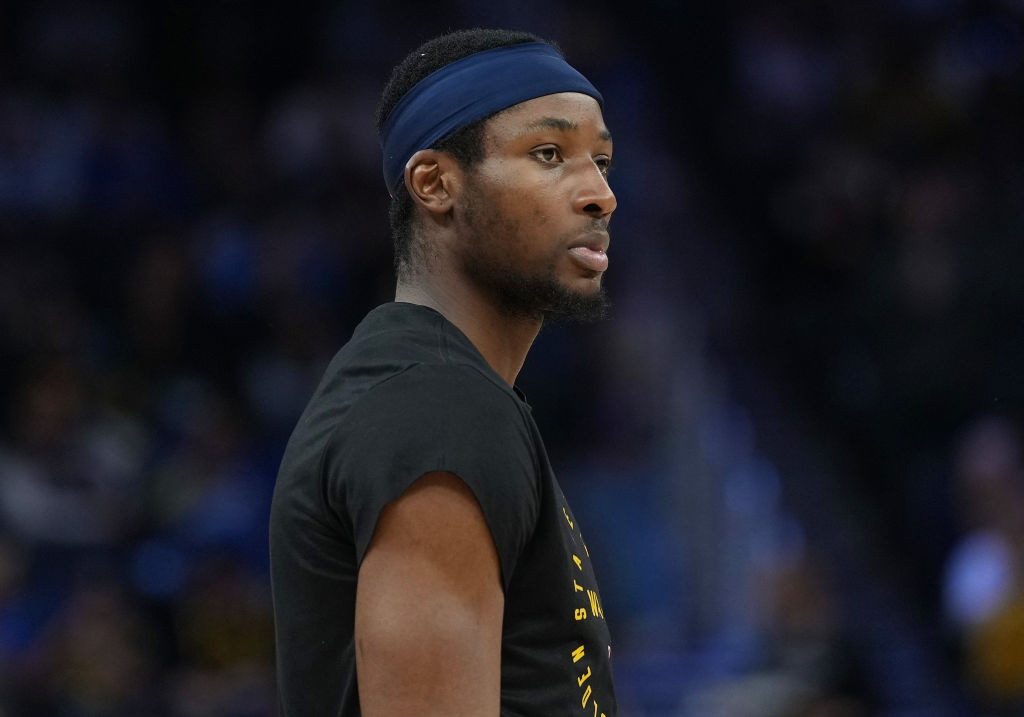 A man with a beard and short hair wears a headband and a black shirt. He's looking intently to the side, with a blurred, dark background behind him.